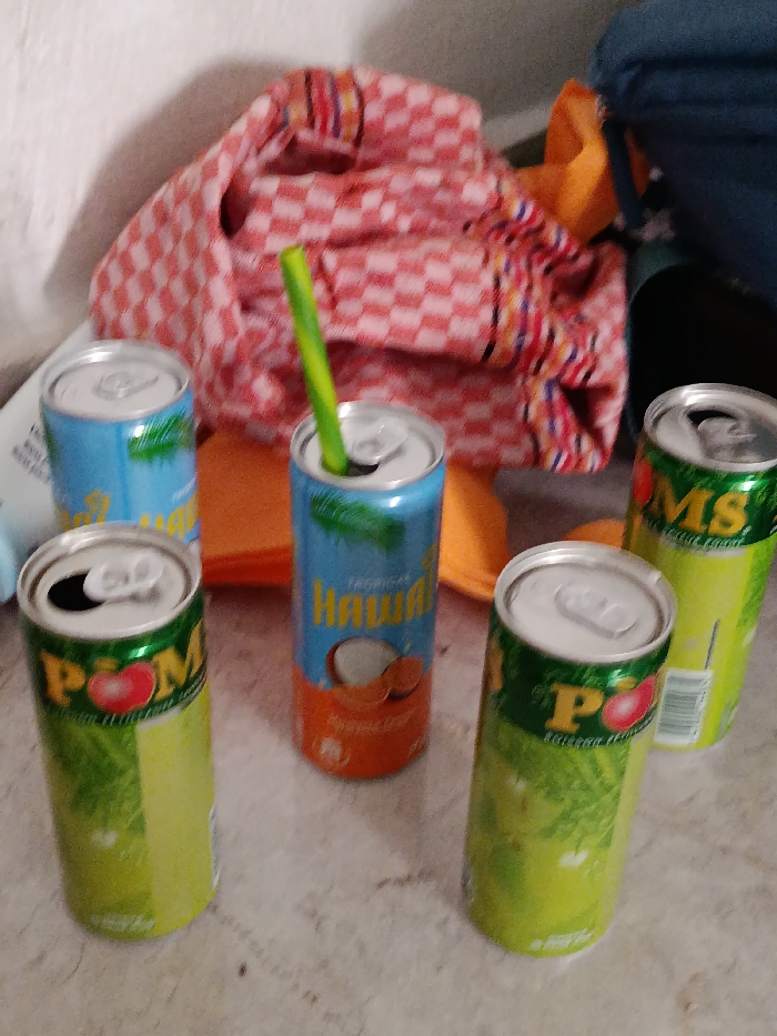 The scene, located in Deroua, Morocco, displays five beverage cans resting on a light-colored, textured surface, possibly a floor. From left to right, an open green can labeled "POME" with a red pomegranate icon is visible, followed by a closed green can of the same brand. Centrally, a tall, blue and orange can, labeled "Tropical Hawai" with a coconut graphic, has its top opened, and a green, flexible straw is inserted into it. To the right, a partially visible green can labeled "MS" with a citrus image appears closed. A fifth blue can is partially visible behind the leftmost "POME" can. In the background, a crumpled red and white checkered fabric is present, partially covering an orange flat object and dark blue fabric. A white bottle is also partly visible on the far left edge of the frame. No individuals are present.