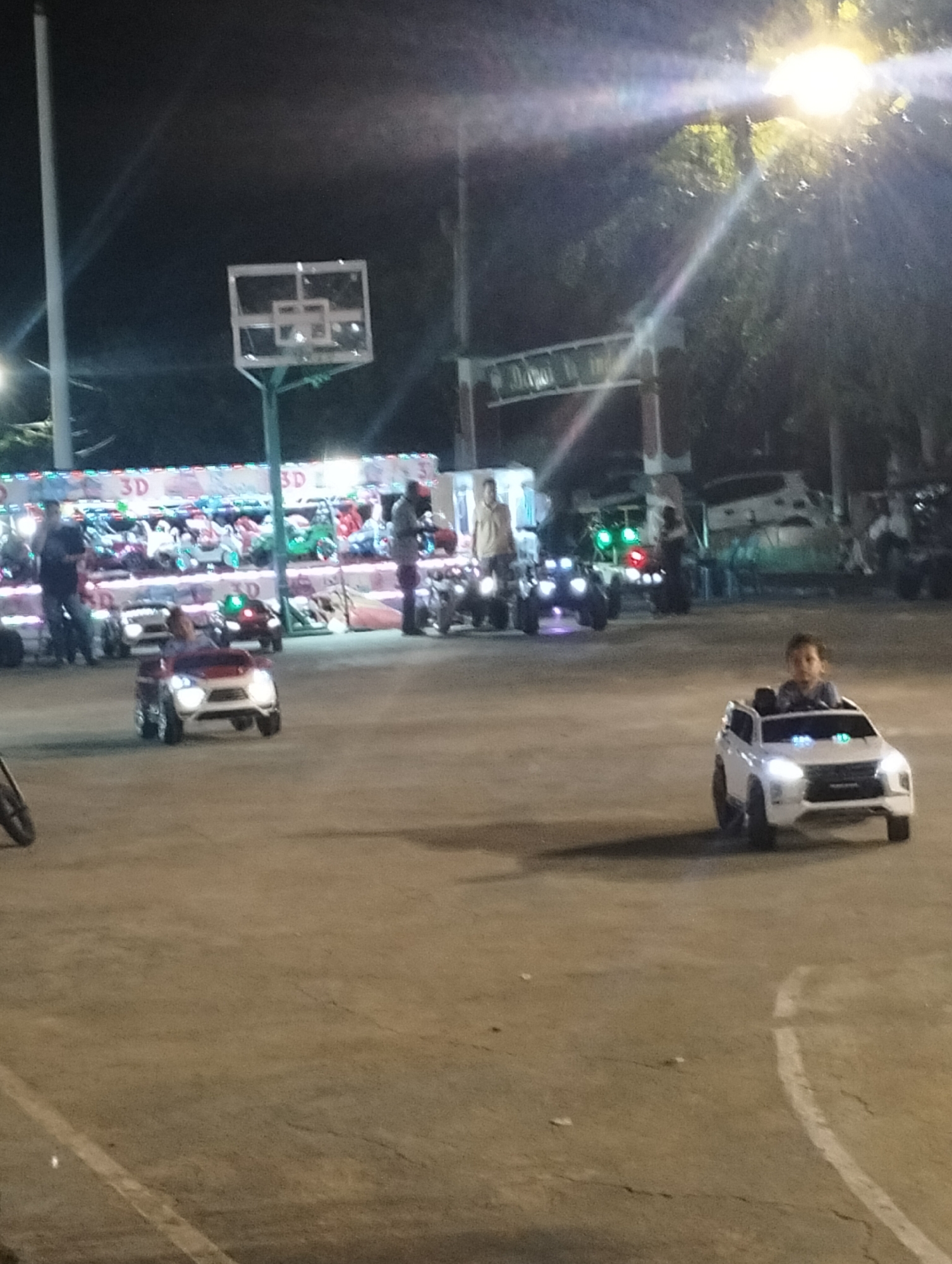 The image shows an amusement area at night in Lhokseumawe, Indonesia. Two children are driving battery-powered toy cars on a paved surface marked with a basketball court line. Other toy cars and people are visible in the background, along with a basketball hoop, fairground ride, and illuminated sign. Overhead streetlights and reflections contribute to the scene's lighting.
