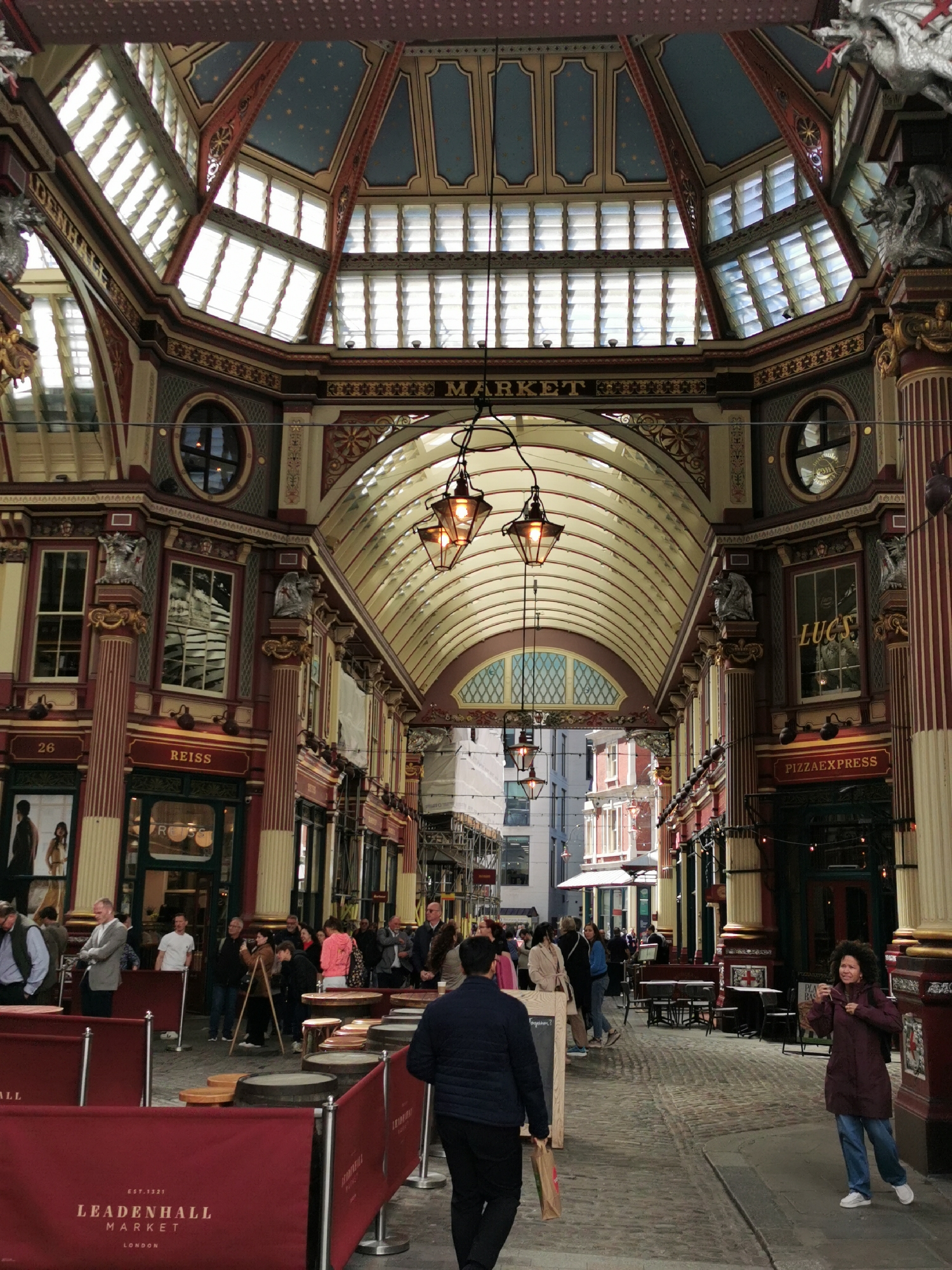 This image captures the vibrant interior of Leadenhall Market in London, United Kingdom, a historic covered market showcasing ornate Victorian architecture. The scene is bustling with numerous people engaged in various activities, suggesting a lively daytime atmosphere.
The market features a grand, high ceiling with intricate dark red, gold, and blue structural elements. Sections of the ceiling are painted with stars and incorporate numerous skylights and arched windows, allowing natural light to illuminate the space. Ornate pillars, some adorned with decorative gargoyles or dragon-like sculptures, line the pedestrianized cobblestone walkway. Hanging lanterns with warm, glowing lights are suspended along the thoroughfare.
On both sides of the pathway are shopfronts and establishments. Visible text includes "REISS," "LUCS," and "PIZZAEXPRESS." Red banners or barriers clearly display "LEADENHALL MARKET EST. 1321 LONDON," indicating its historical significance and location. Within these sectioned areas, wooden barrels serve as tables, hinting at dining or casual gathering spots.
A diverse group of people is seen walking, standing, and conversing throughout the market. A woman in a dark purple coat, blue jeans, and white trainers is notably smiling as she walks on the right side of the frame. The overall impression is one of a busy, yet relaxed, urban market environment.
