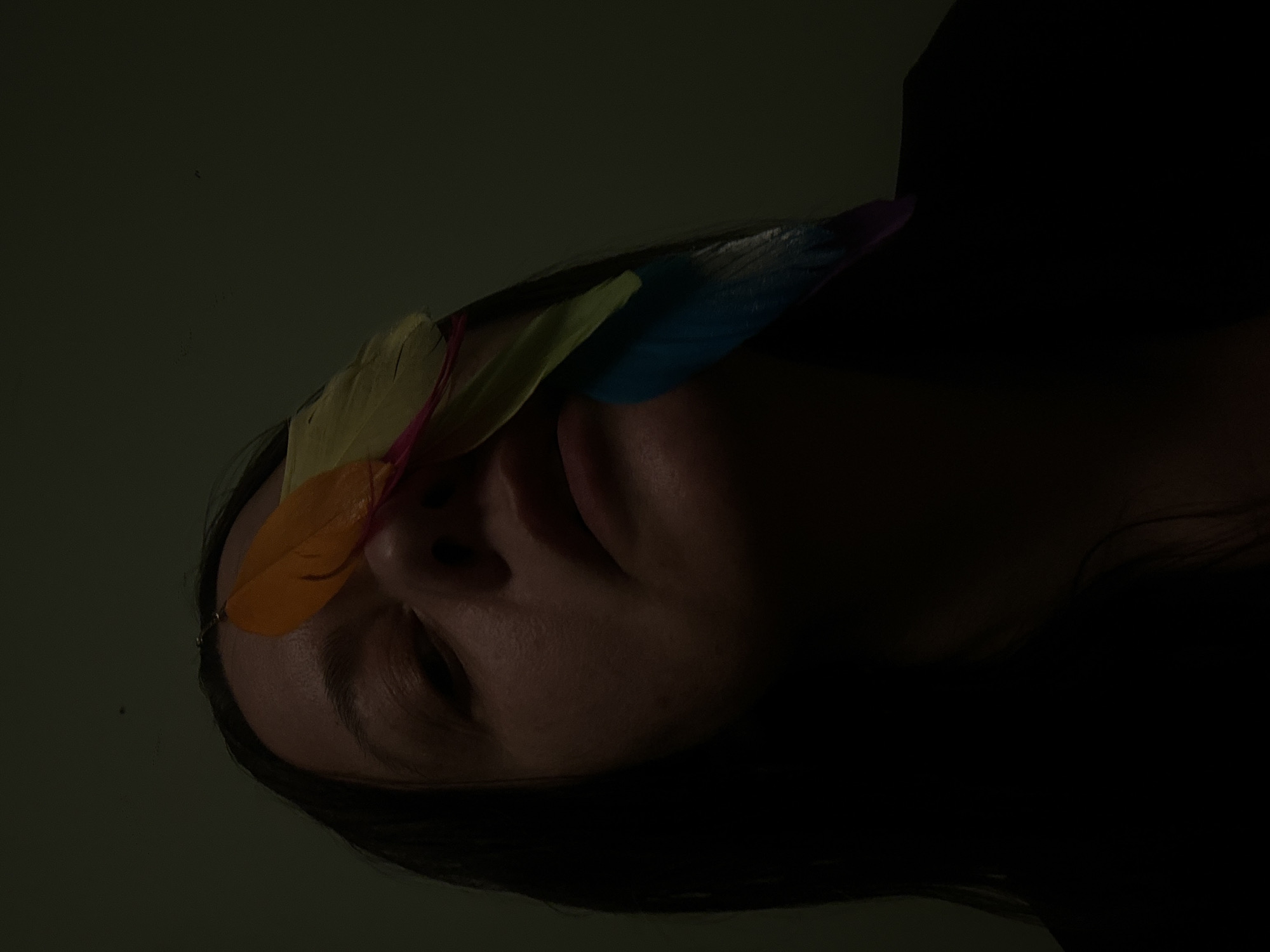 The image presents a dimly lit scene featuring a person lying down with several colorful feathers resting on their forehead and nose area. The feathers appear to be orange, yellow, and blue. The person has dark hair and their eyes are partially closed. The background is a muted, dark green. The lighting is poor, obscuring details, especially in the person's hair and upper body. The setting is not identifiable as Riga, Latvia from the visual information available.