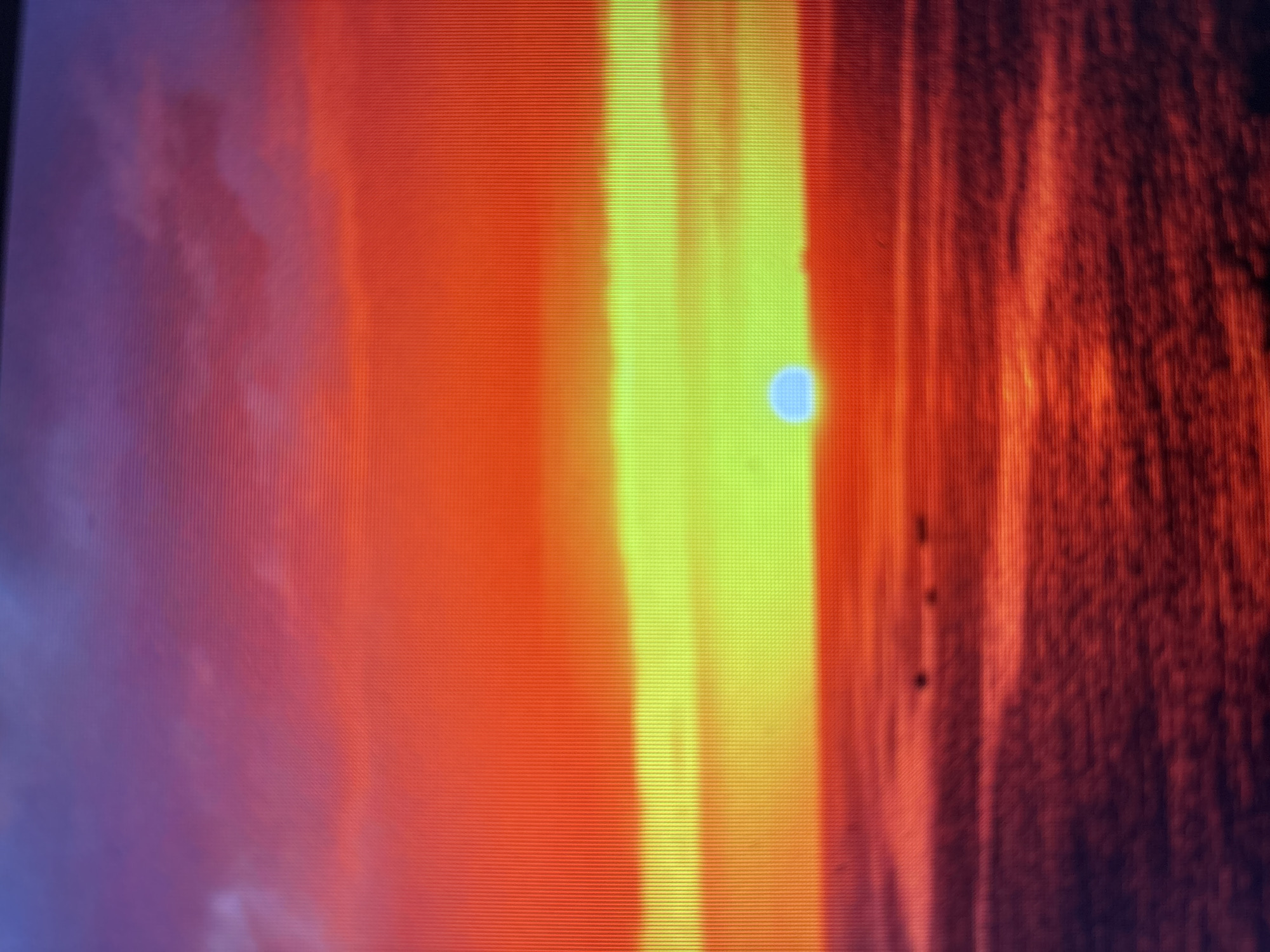 The image shows a blurred, close-up view of a screen displaying an abstract, vertical composition of warm colors. On the left, there is a purple and red area, transitioning into a brighter orange. A vertical band of yellow runs through the center, with a small blue spot visible on it. To the right of the yellow, there is a darker reddish-brown area that has some vertical streaking. The screen's pixel structure is clearly visible. The location in Durmersheim, Germany is unknown.