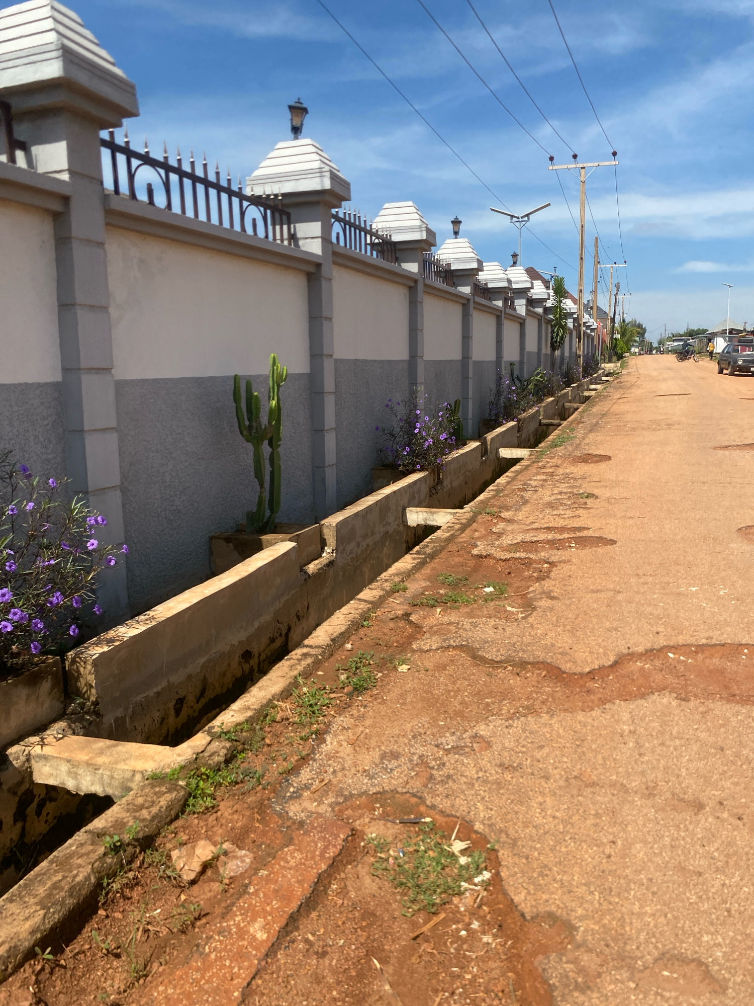 A street in Barden Yana, Nigeria, is depicted with a drainage ditch running alongside a tall, decorative wall. The wall features square pillars topped with pyramid-shaped caps and ornate metal fencing. Several plants, including a cactus and purple flowers, are positioned within planters along the base of the wall. The street itself is unpaved and made of reddish-brown earth, with some signs of wear and minor puddles. Power lines are visible overhead, supported by wooden poles. In the distance, a few vehicles and people are present, indicating a residential or semi-urban area. The sky is blue with scattered white clouds.