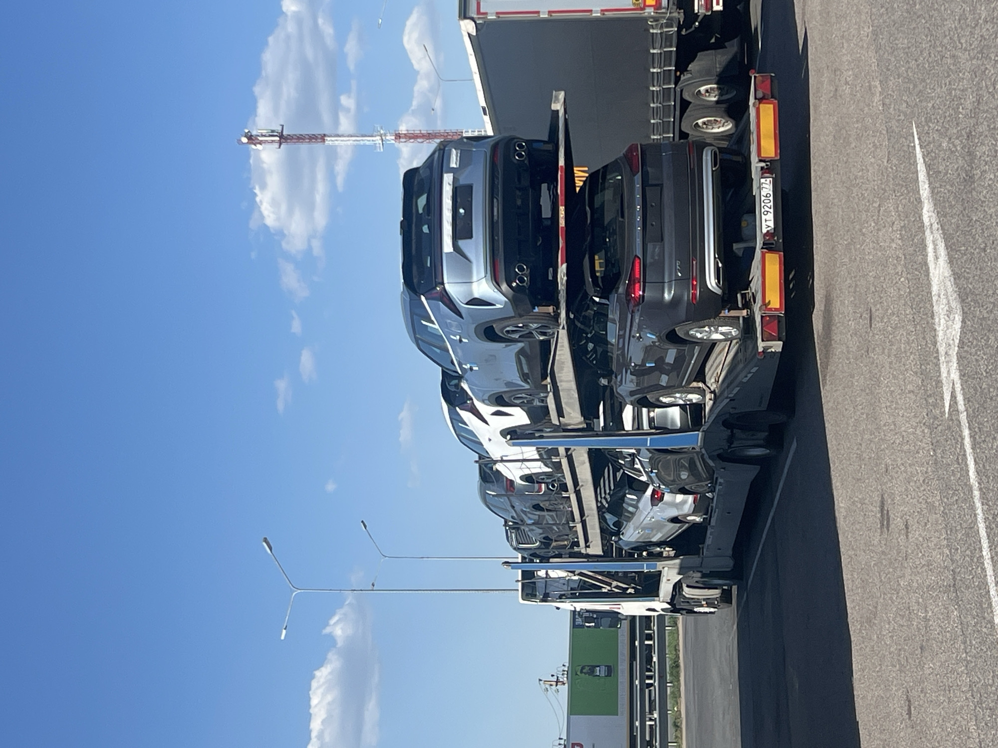 The image shows a car carrier truck loaded with new cars, likely in transit, parked on a paved surface alongside a road. The sky is blue with scattered white clouds. A communication tower is visible in the background. The location is Казань, Russia, identifiable by the license plate "YT 9206 77" on the trailer, which corresponds to Russian vehicle registration codes. Streetlights are also present along the road. There is a large green and white building sign near the truck.
