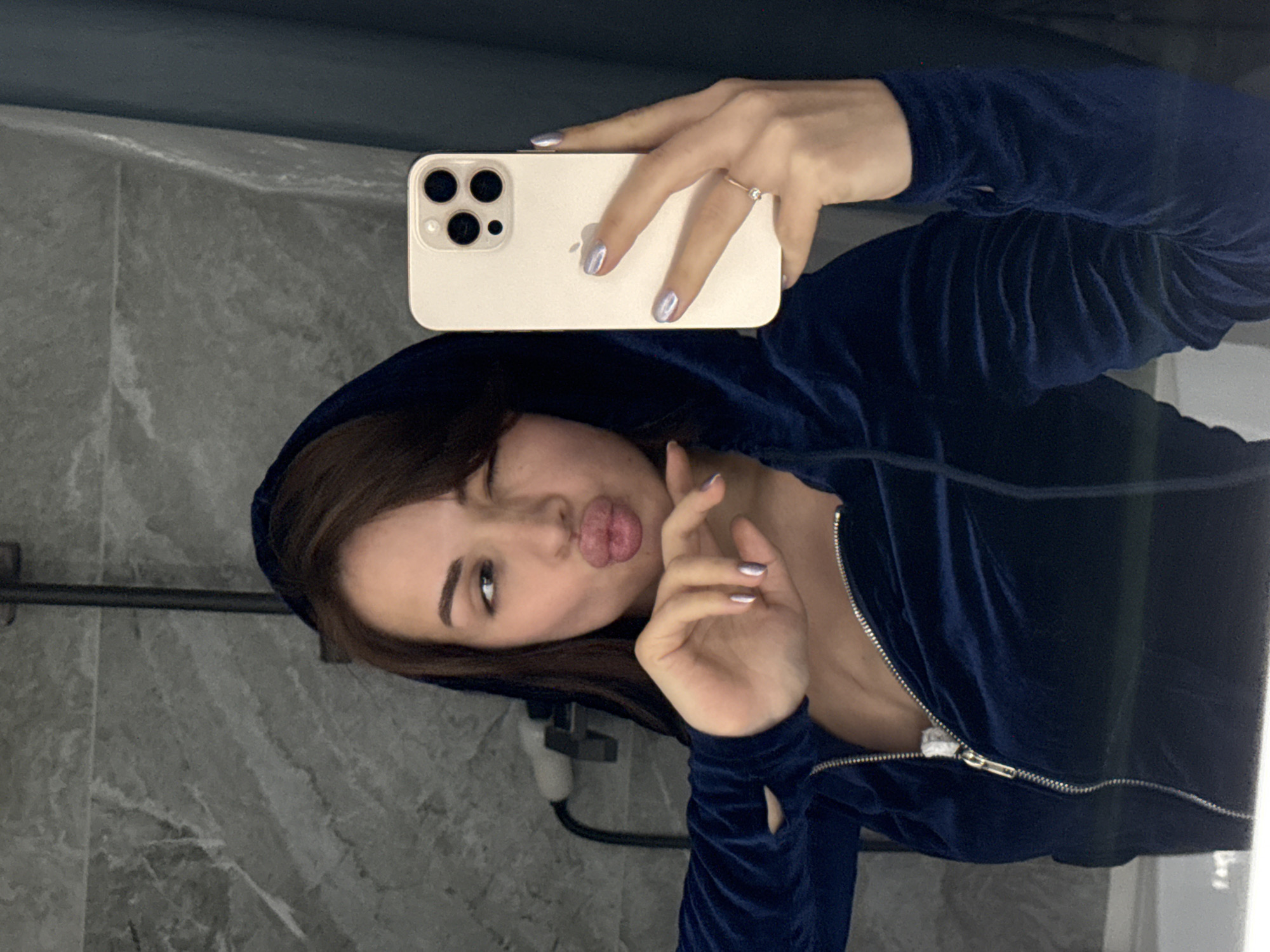 A woman is taking a selfie in a bathroom. She is wearing a navy blue velvet zip-up hoodie. She has light-colored fingernails and a ring on her ring finger. The background appears to be a grey tile wall. The phone she is holding is a light color with multiple lenses. The location is Лобаново, Russia.
