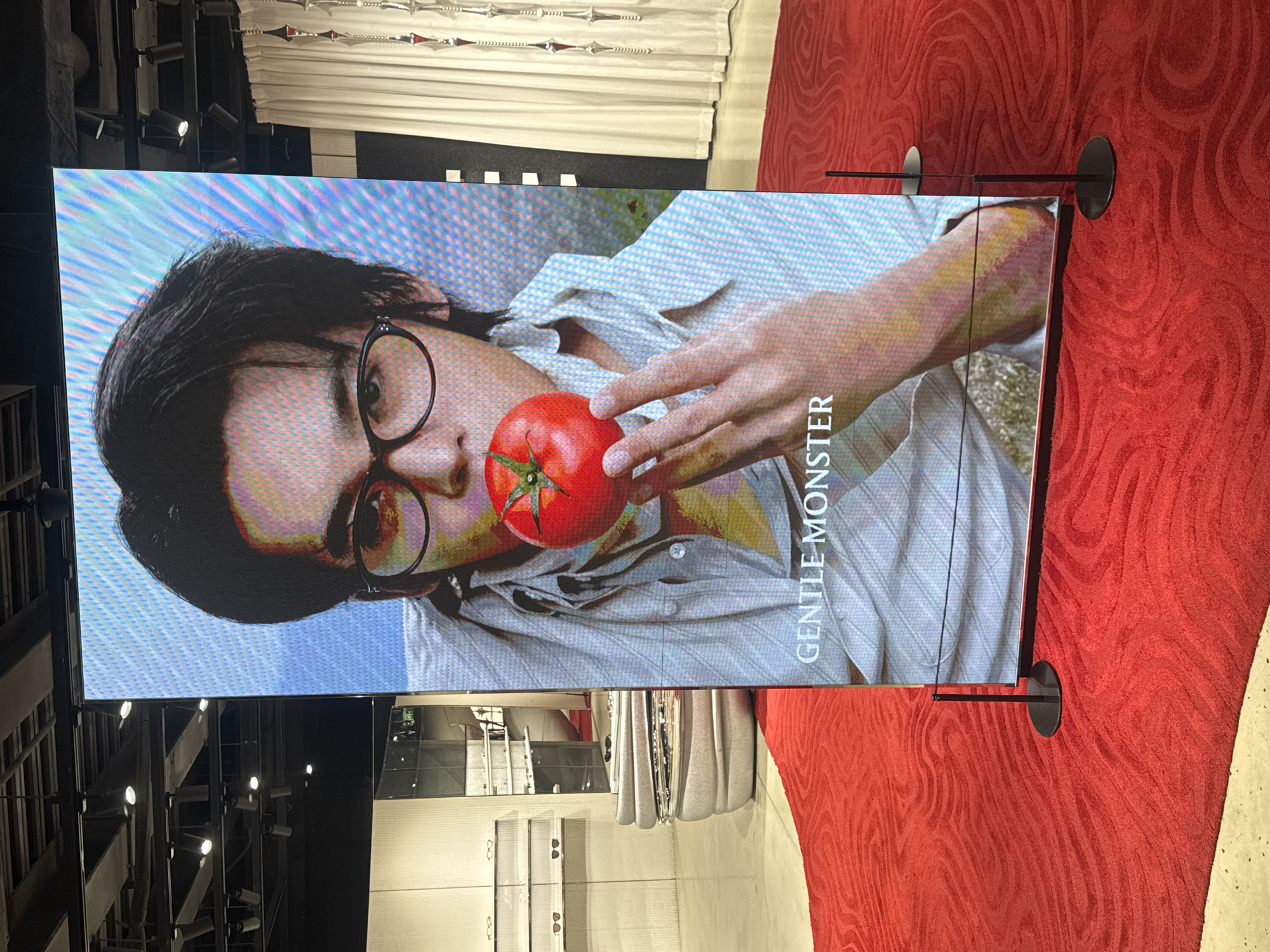 A large, vertical LED screen displays a close-up of a person holding a tomato to their face, with the text "GENTLE MONSTER" at the bottom. The screen is positioned on a red carpet with a textured, wave-like pattern. In the background, to the left of the screen, is a retail store interior with shelves displaying eyeglasses, a modern-looking display case, and a mirrored surface. To the right of the screen, white curtains hang, and decorative metallic elements are visible. The overall environment suggests a retail space in Shanghai, China.