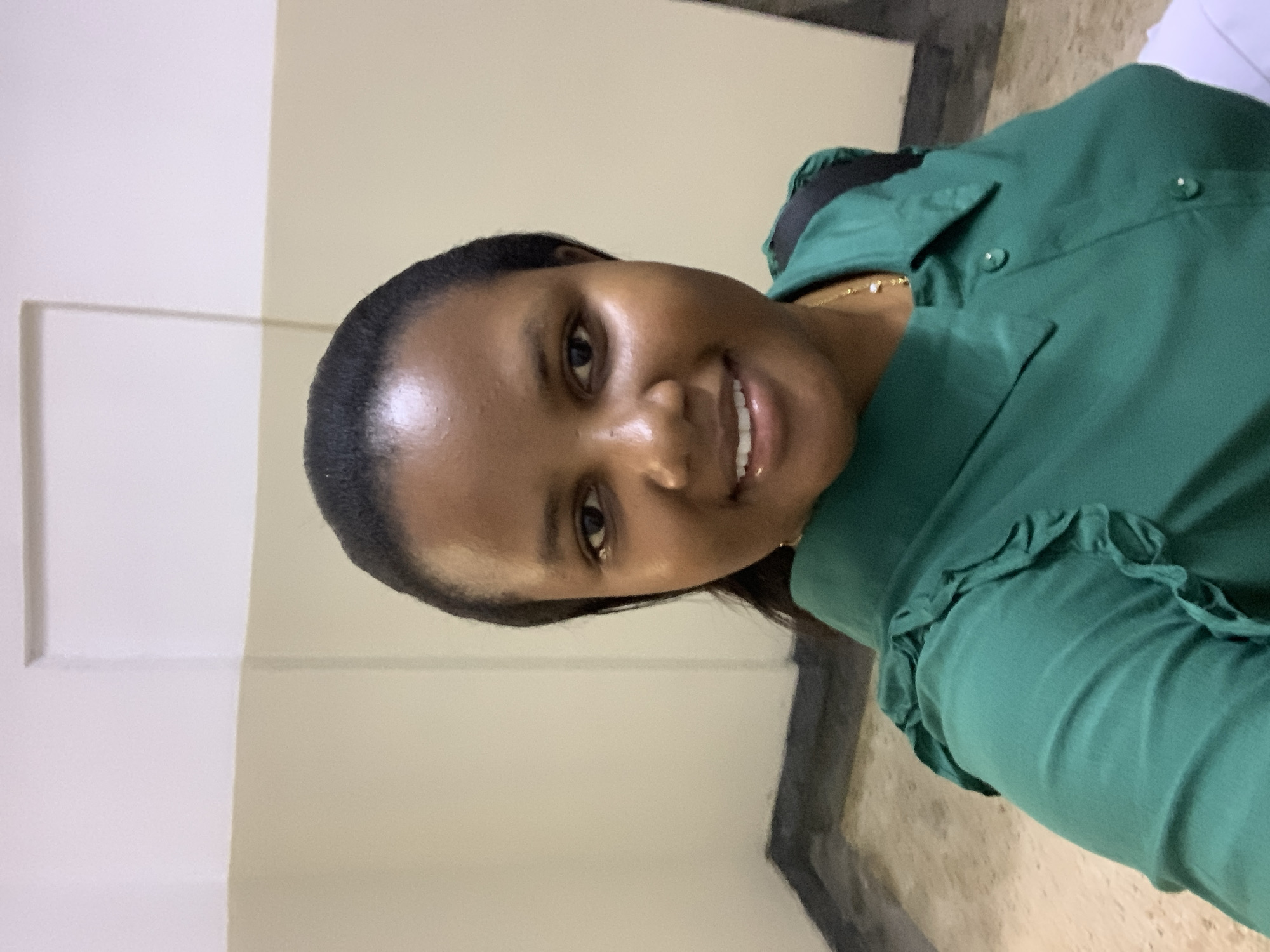 A woman with dark hair and brown skin is smiling at the camera. She is wearing a green shirt with ruffles on the sleeves. The background is a wall with two different shades of beige. The location is unknown and cannot be determined from the image.