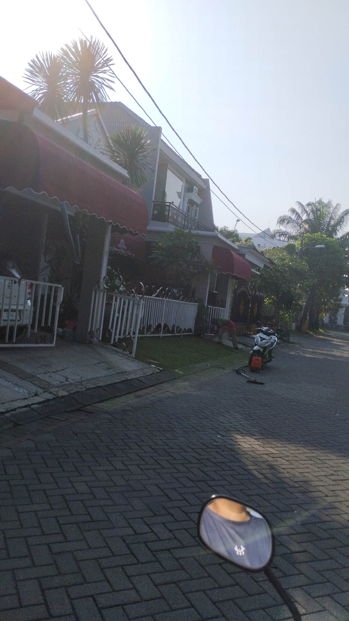 In the city of Sidodadi, Indonesia, a residential street is captured on a clear day. The foreground is dominated by a paved walkway, with a motorcycle side mirror reflecting the neck and shoulder of a person wearing a grey t-shirt with a white logo. Beyond the walkway, a white picket fence borders a manicured lawn in front of a modern multi-story house with red awnings over the windows. A person in a green shirt is bent over near the lawn, possibly tending to it. A white pickup truck is parked in front of the house. A scooter is parked on the street. Electrical wires are visible crisscrossing the sky above the houses and trees. Palm trees and other lush vegetation surround the property, suggesting a tropical or subtropical climate. The scene conveys a sense of peaceful, suburban life in Indonesia.
