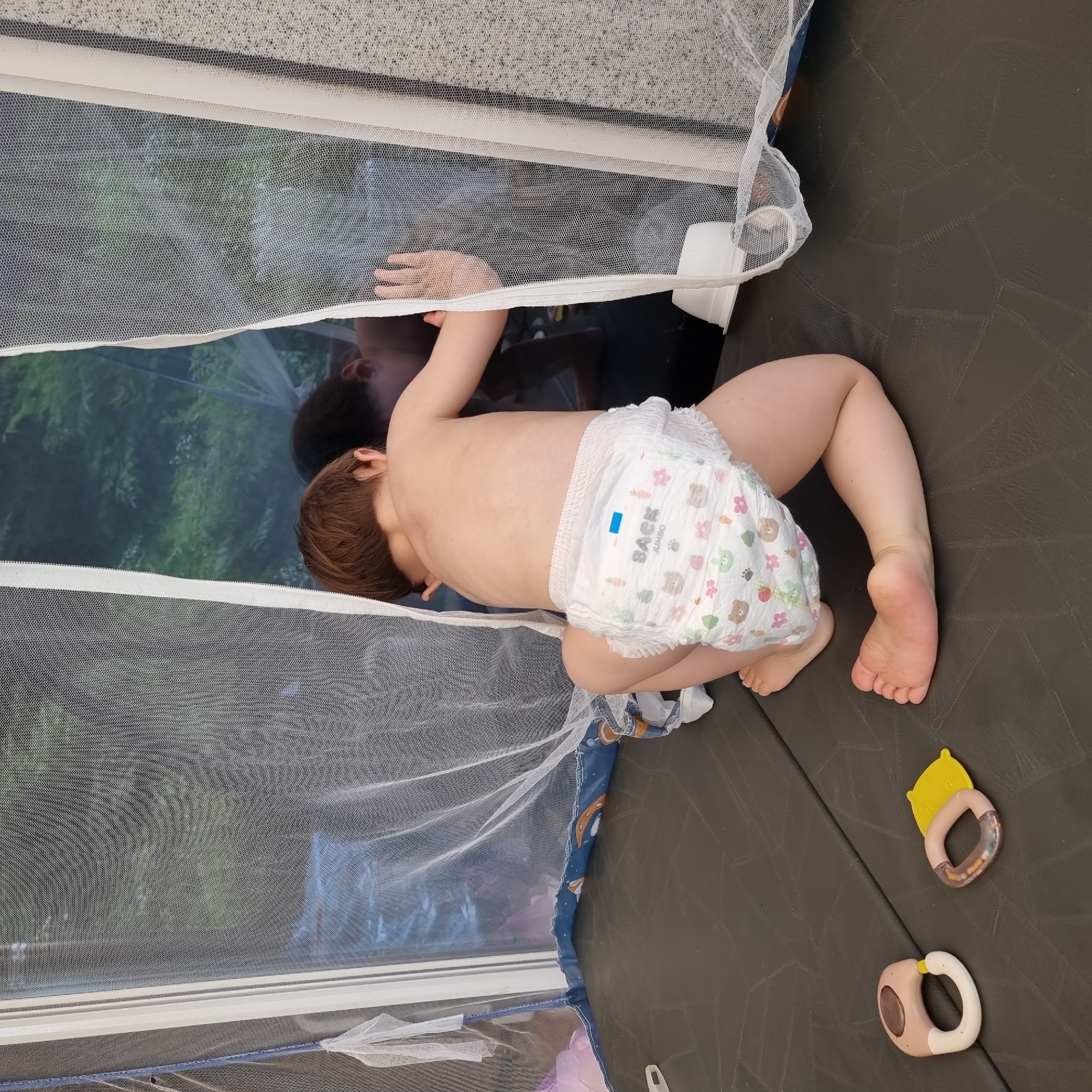 A young child, wearing a diaper adorned with cartoon animal prints, is seen from behind. The child is on their hands and knees on a dark, textured surface, likely a mat. They are looking out through a mesh window or tent opening, with their right hand pressed against the mesh. The child's reflection is visible in the dark surface beyond the mesh. Two baby rattles are visible on the mat in the foreground. The scene appears to be indoors or in a sheltered outdoor space, given the presence of the mesh tent-like structure. The specific location within Dunnae-myeon, South Korea cannot be determined from the image alone.