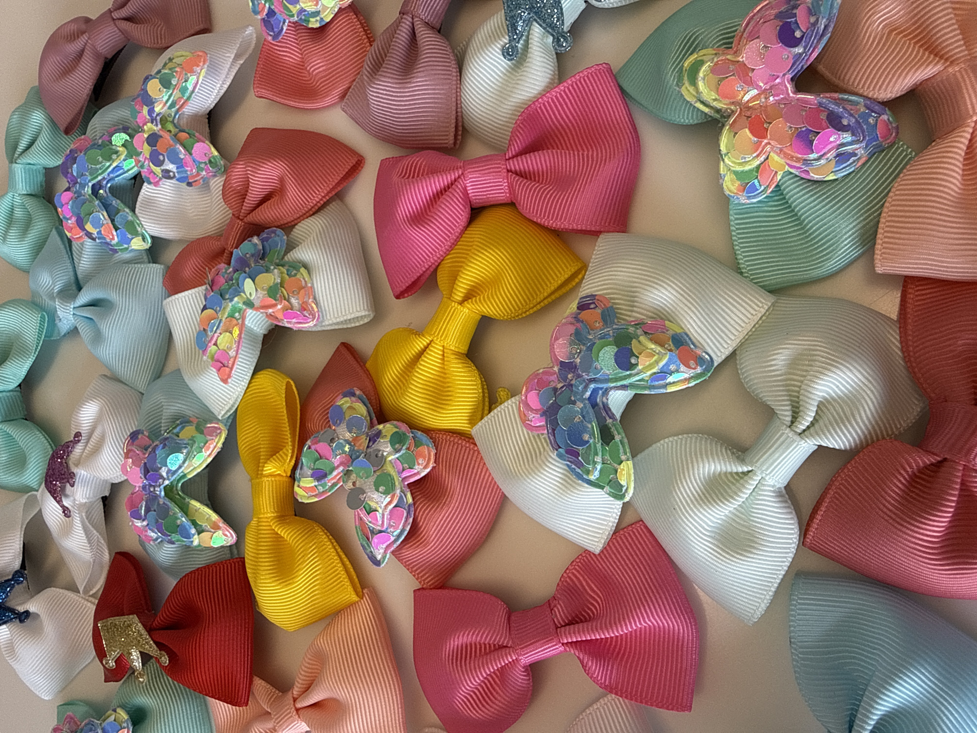The image displays an assortment of colorful hair bows scattered across a flat surface. The bows are predominantly made of grosgrain ribbon in shades of pink, yellow, white, red, blue, and green. Some bows feature decorative embellishments such as sequins shaped like butterflies or small crowns. The surface is a solid, light-colored material. No people are visible. The location is not identifiable from the image.