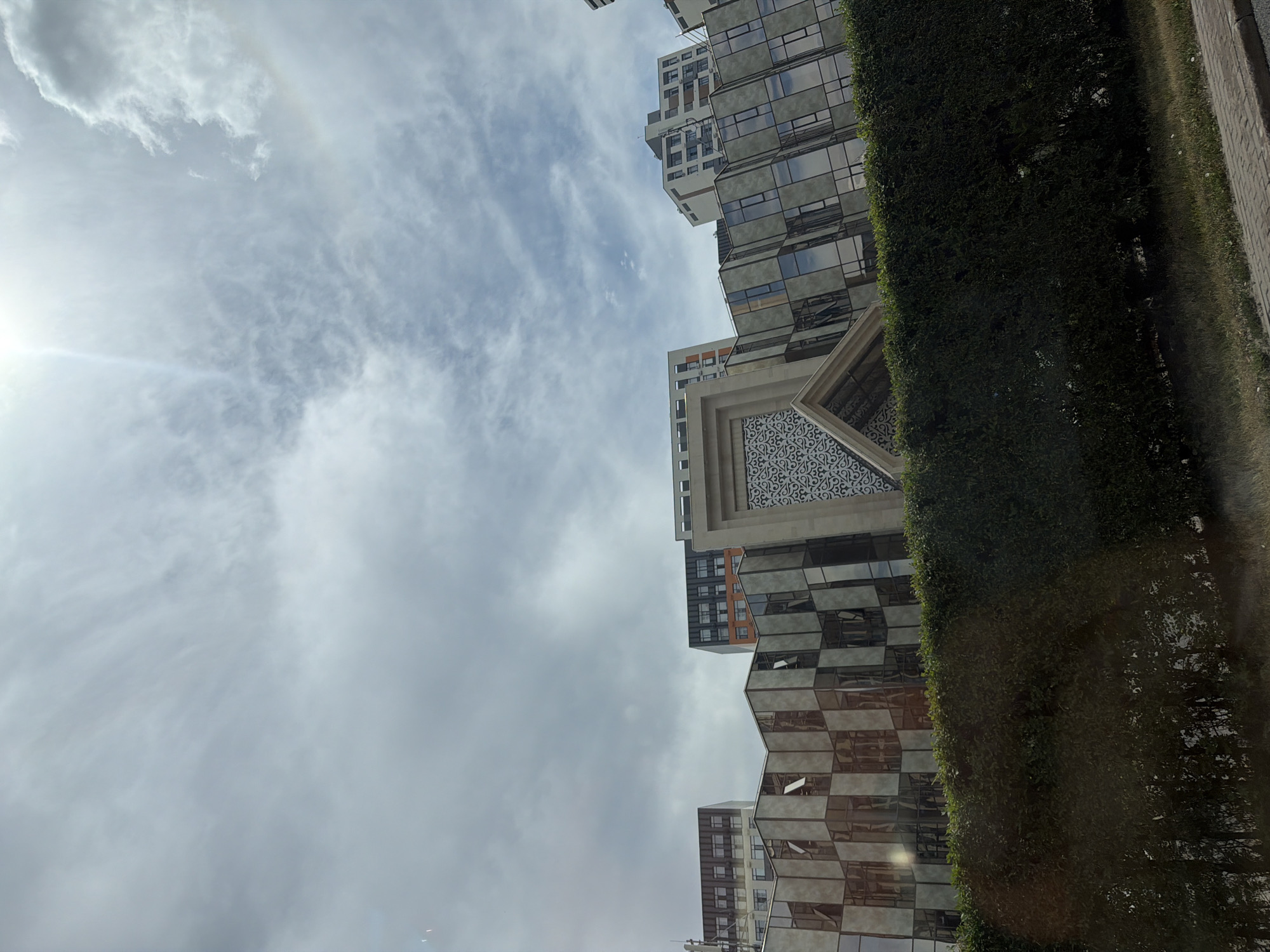 This media file shows an upward-angled view of modern buildings against a cloudy sky. The buildings have a contemporary architectural style, featuring large glass windows and geometric shapes. One building displays an ornate, patterned facade. A tall, dense green hedge runs alongside the buildings. The sun is partially visible on the left, creating a slight halo effect in the sky, suggesting it is daytime. The weather appears partly cloudy, with a mix of bright sky and overcast clouds. The scene evokes a sense of urban development and modern city living. The image does not contain any discernible text.