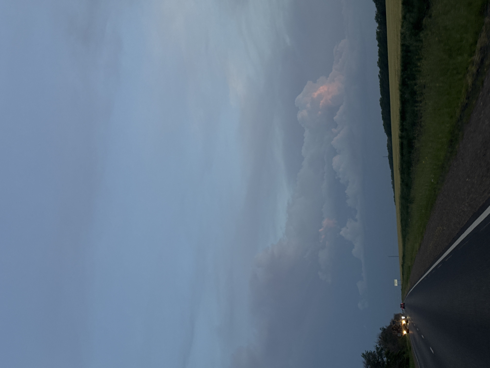 The image shows a road near Beysug, Russia. The sky is cloudy, with a large cloud formation in the center. A field and some trees line the right side of the road. A vehicle with illuminated headlights is visible further down the road. The road is paved with a white line marking the edge.