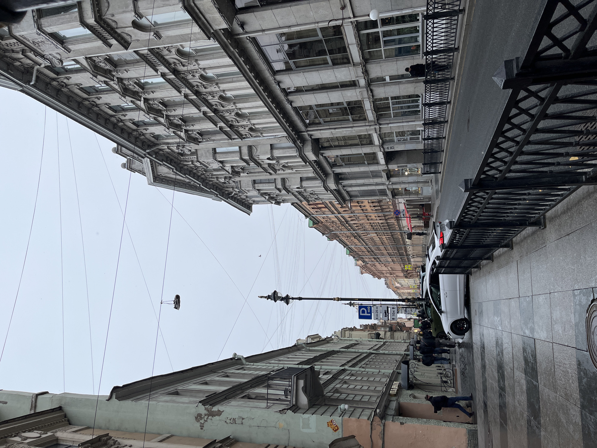 An urban street scene unfolds under an overcast sky, framed by tall, multi-story buildings on both sides. The perspective looks down the length of the street, with buildings receding into the distance.
On the left side, an older building features a facade with ornate detailing and areas of peeling, pale green paint on its lower levels. Numerous thin wires crisscross the sky above this building and across the street. Notably, a single shoe hangs suspended from one of these overhead wires in the upper left quadrant of the image.
On the right, another large building, predominantly grey, lines the street. A black metal fence with decorative patterns runs along the sidewalk, separating it from the roadway. The street itself is paved with asphalt, and several cars are parked along the right curb, including a prominent white sedan.
People are visible on both sidewalks, walking and standing. A group of individuals is clustered near the parked cars on the right, and a few more are further down the street. In the mid-ground, a tall, decorative black street lamp post stands.
The sky is a uniform, dim grey, indicating an overcast day with no direct sunlight. The overall lighting is soft and muted. On the right sidewalk, a circular manhole cover is visible on the grey pavement.
Visible text includes a blue parking sign with a "P" symbol, displaying "8:00 - 20:00" (likely parking hours) and "↑ 10м ↑". On the lower left facade of the older building, a sign with Cyrillic script is partially visible, appearing to read "ОТДЕЛЕНКИ". The architectural style and visible text suggest a European city, possibly in Russia.