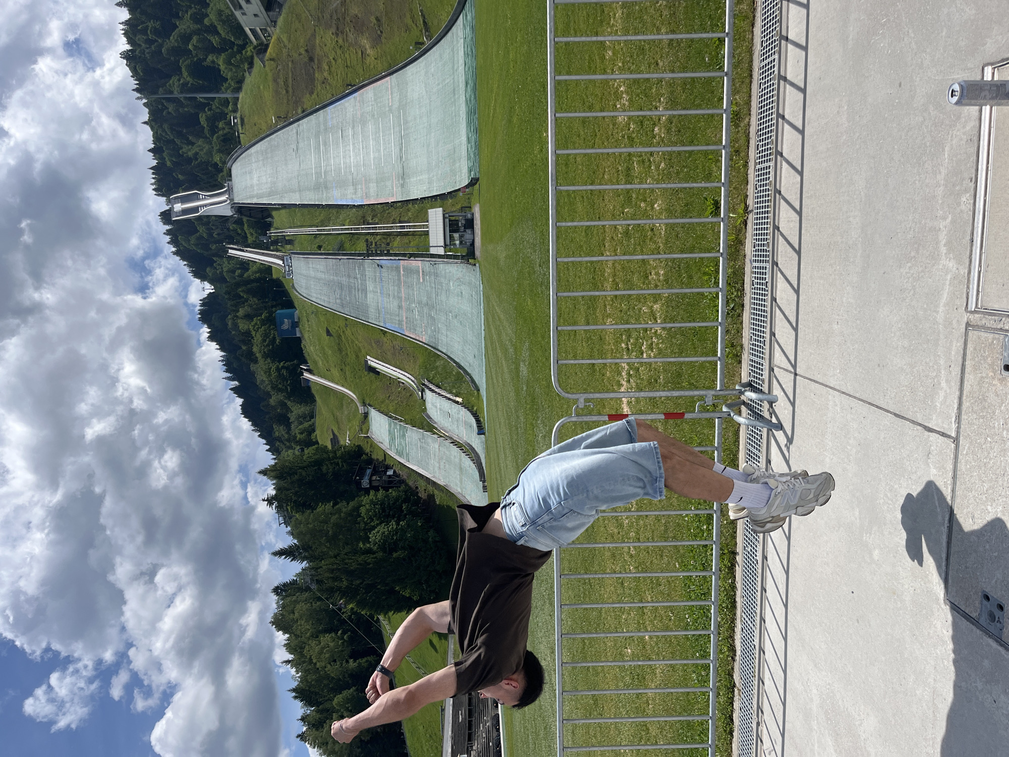 A male individual is captured mid-air, performing an acrobatic backflip or similar inverted jump, in front of the Olympiaschanze ski jump complex in Garmisch-Partenkirchen, Germany. He is wearing a dark brown t-shirt, light blue denim shorts, white socks, and white sneakers, with his arms extended upwards and backwards, and his body arched. His head is tilted back, facing towards the ground. The ski jump complex in the background features multiple large ramps covered in green matting, indicative of summer training surfaces, ascending a steep, tree-covered mountain. The slopes are flanked by dense coniferous forest under a partially cloudy blue sky. A silver metal barrier fence, composed of vertical bars, stretches across the mid-ground, separating a grassy area from the concrete pavement in the immediate foreground where the individual is jumping. Shadows of the fence are cast on the concrete surface, which also features drainage grates.