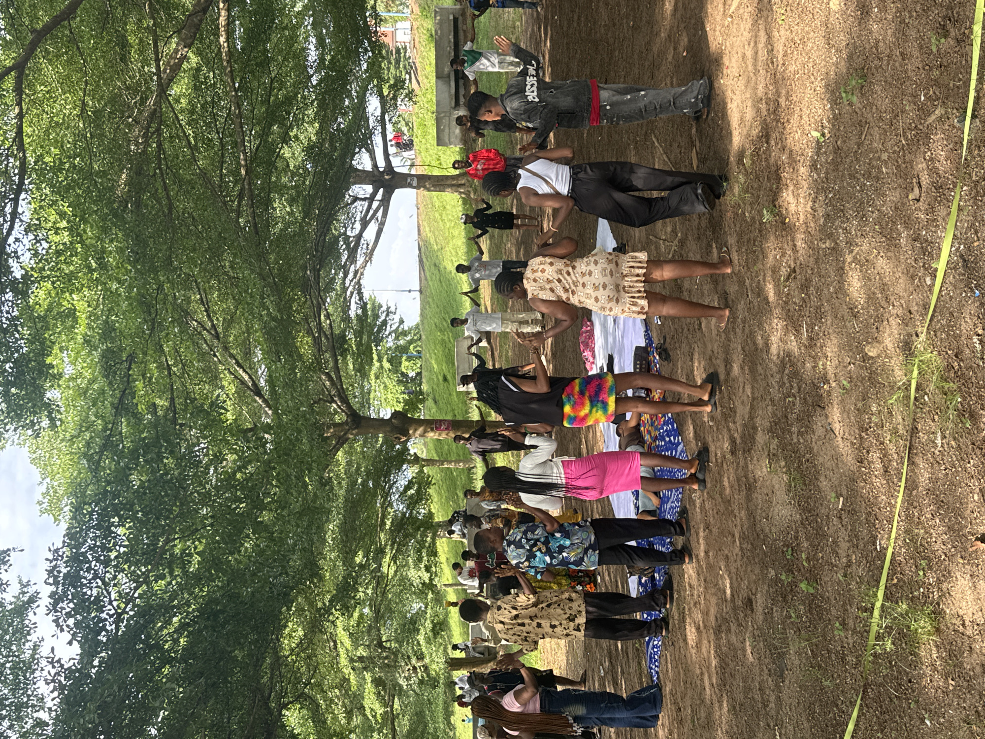 A group of people stand in rows beneath a large tree in Ihiagwa, Nigeria. They appear to be engaged in a group activity, possibly a dance or exercise routine. The ground is dirt and partially covered in grass and tree shadows. A yellow ribbon lies on the ground to the right. The background shows other trees and what appears to be a structure in the distance.