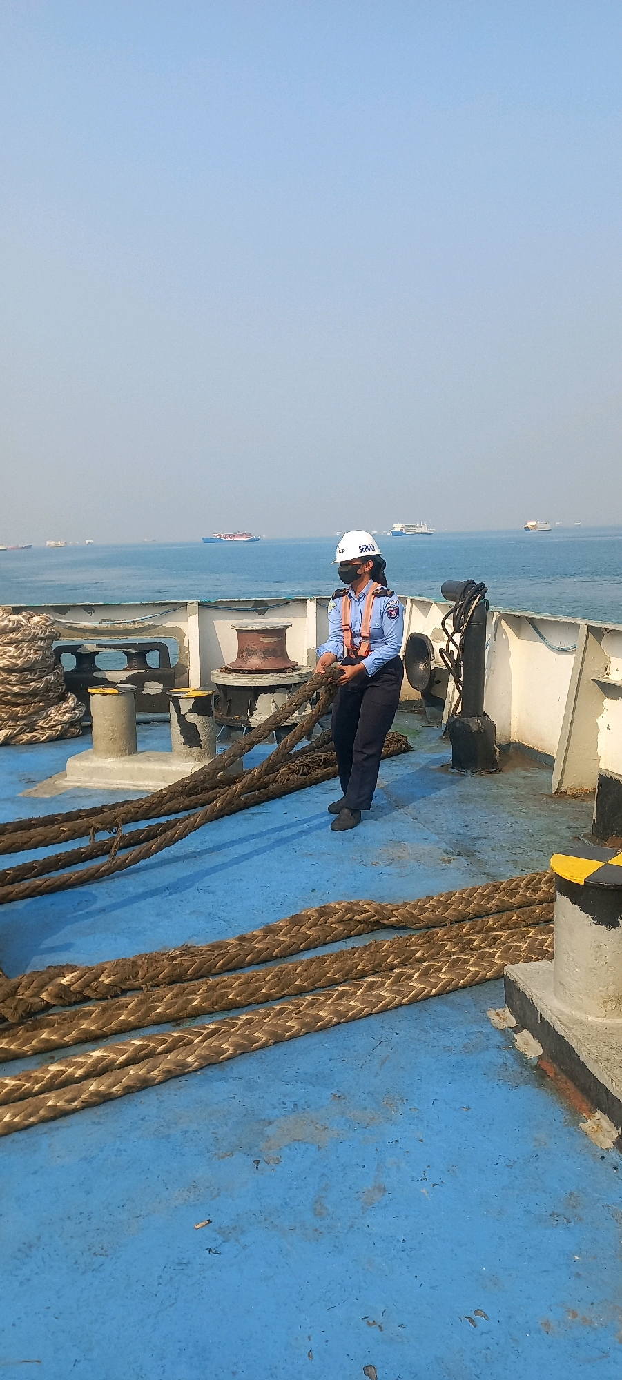 The image shows a person in a hard hat and safety vest on the deck of a ship. The person is holding a thick rope. There are other ropes coiled on the deck, and some winches are visible. In the background, the ocean can be seen with several ships in the distance. The sky is clear and blue. The scene appears to be in a port or at sea. The location of Cilegon, Indonesia, is not explicitly indicated by any visual cues in the image.