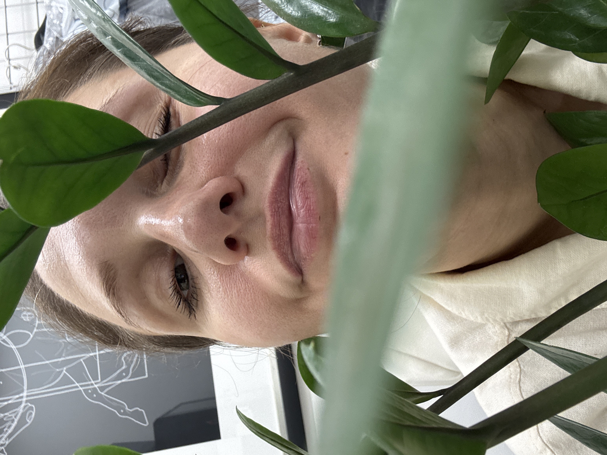 The image presents a close-up, angled portrait of a person, framed and partially obscured by a large, leafy green plant. The photograph is oriented with the subject's head tilted to the right, implying a 90-degree counter-clockwise rotation from a typical upright view. The subject is a female with fair skin, light brown hair, and visible dark eyelashes. Her face, illuminated by soft, natural light, displays a gentle, closed-mouth smile and a relaxed expression, with her visible eye gazing slightly upwards towards the viewer. A natural sheen is apparent on her skin.
Dark green stems and glossy leaves of a plant, resembling a ZZ plant, dominate the foreground and midground. These botanical elements intricately frame the subject's face, with some leaves partially covering her left eye and nose, adding a layer of depth and intimacy to the composition.
In the background, to the left of the subject's head, is a dark surface adorned with white line-art drawings that appear to be anatomical or technical diagrams, possibly depicting skeletal structures or machinery. To her right, a light-colored, textured fabric, likely part of her attire (an off-white or cream garment), is visible behind the plant stems. A plain white surface is also seen behind her head.
The overall impression is one of calm and natural beauty, captured in a casual indoor setting. There is no discernible text within the image. The contextual location for this media file is Dubai, United Arab Emirates.