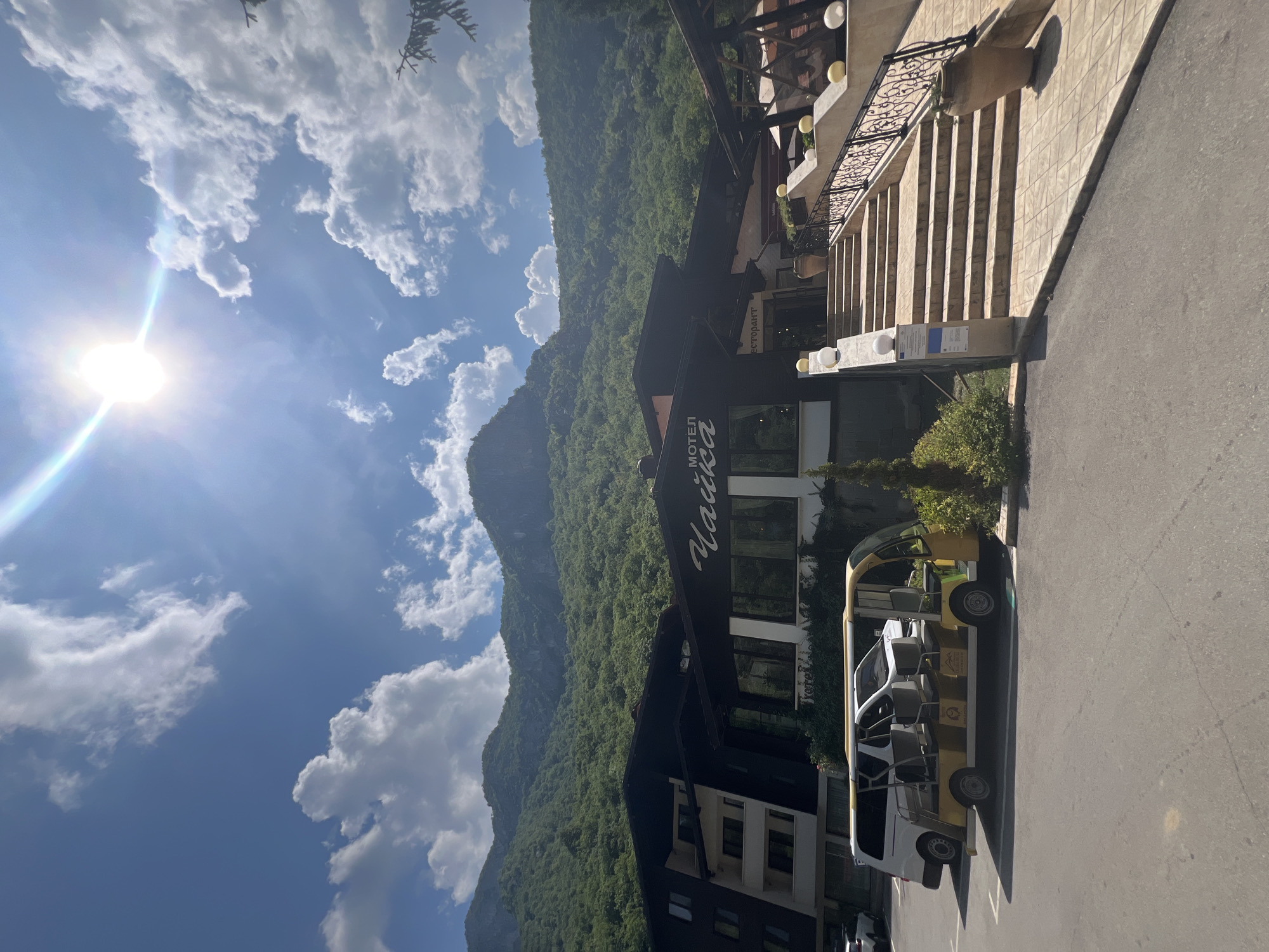The image shows a motel with a restaurant, nestled at the base of a lush, green mountain range under a bright, sunny sky with scattered clouds. The motel, labeled "MOTEL ЧАЙКА" (Motel Chayka), features a dark, sloping roof and a white facade. A set of stone stairs leads up to the entrance of the restaurant, which is decorated with decorative lighting and potted plants. In front of the motel, several vehicles are parked, including a white van and a yellow open-air shuttle vehicle. The surrounding environment is natural, with trees and vegetation visible on the mountain slopes. The image appears to be taken in the city of Vratsa, Bulgaria, a region known for its natural beauty and mountainous terrain. There are no people visible in the image.