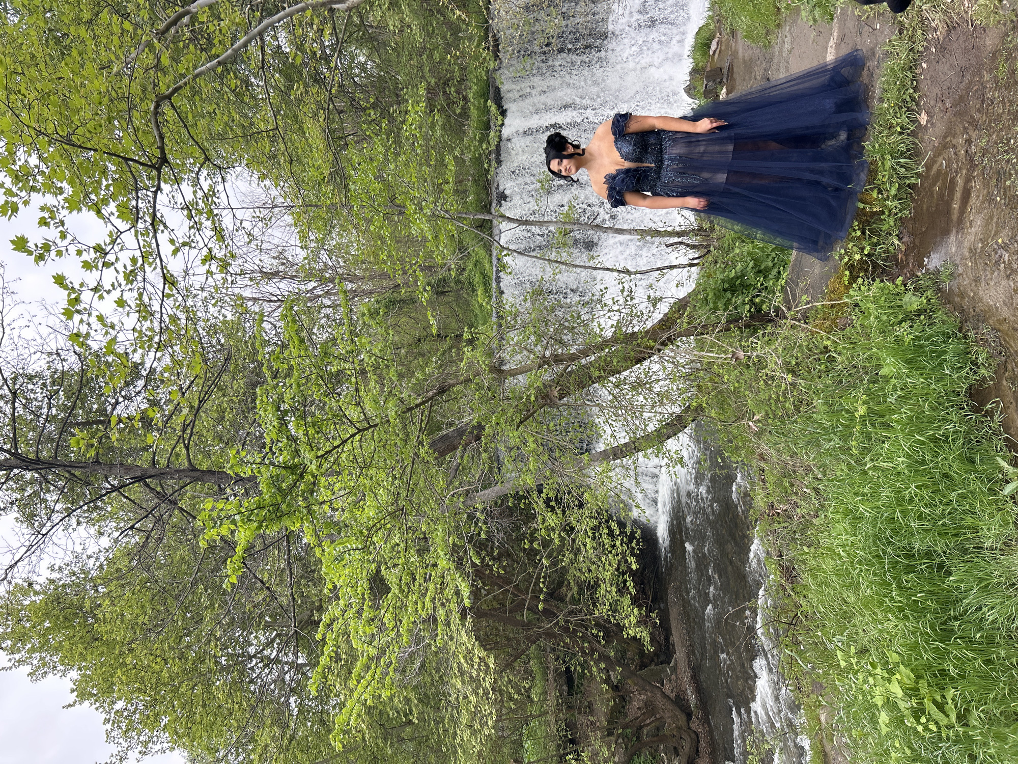 A woman in a dark blue gown stands on a bank next to a waterfall. Her hair is styled up. The scene includes a cascading waterfall, a rocky stream bed, and lush green vegetation with trees framing the view. The location is Parvenets, Bulgaria.