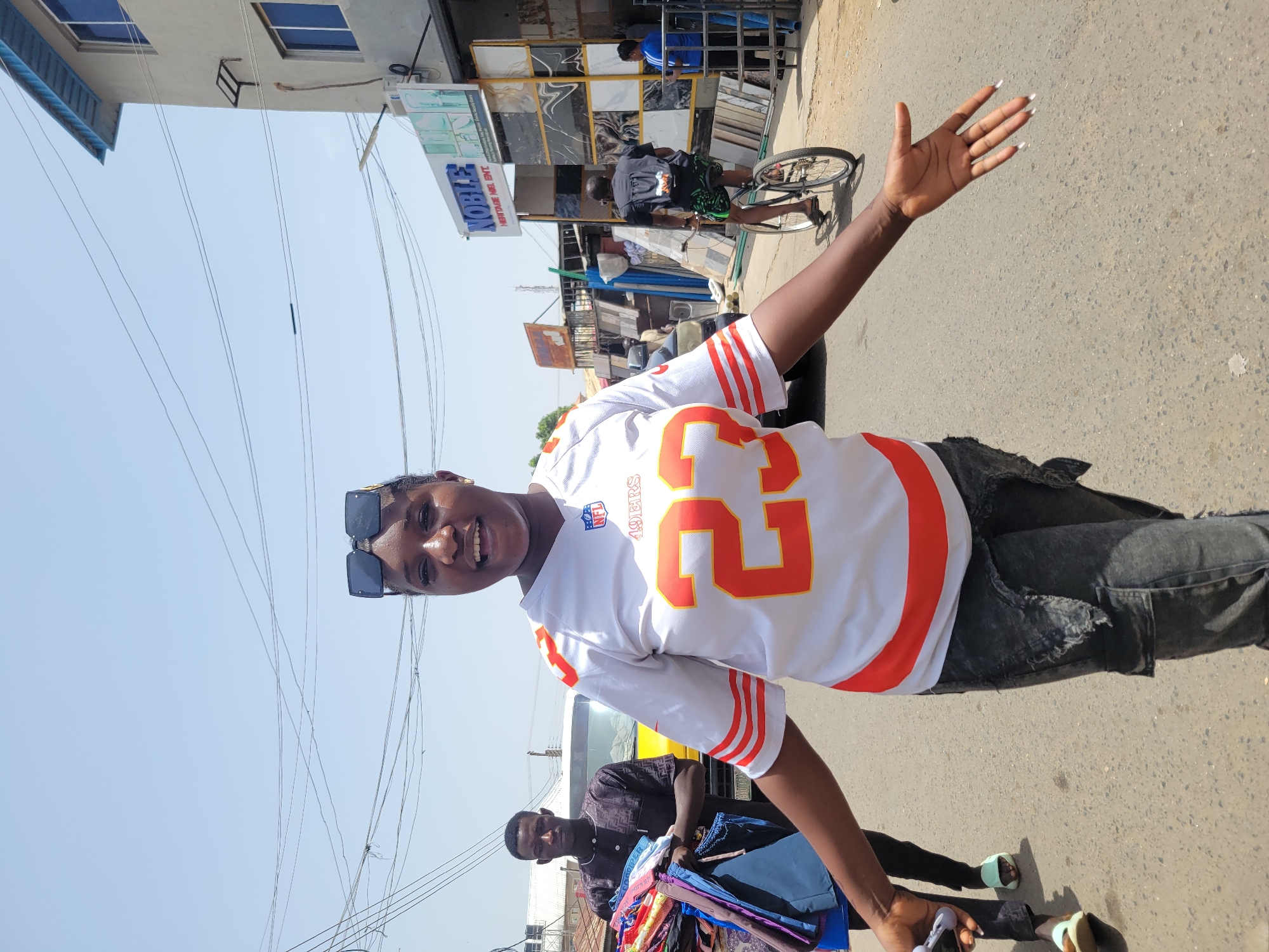 A young woman wearing a white and orange jersey with the number 22 stands on a paved street in Jos, Nigeria. She wears sunglasses atop her head and smiles with one arm raised. Beside her, a man carries a stack of colorful fabrics. In the background, a cyclist passes in front of a building with the sign "NOBLE HERITAGE NIGERIA ENT." A yellow vehicle is partially visible behind the woman. Overhead, a network of electrical wires crisscrosses the sky.