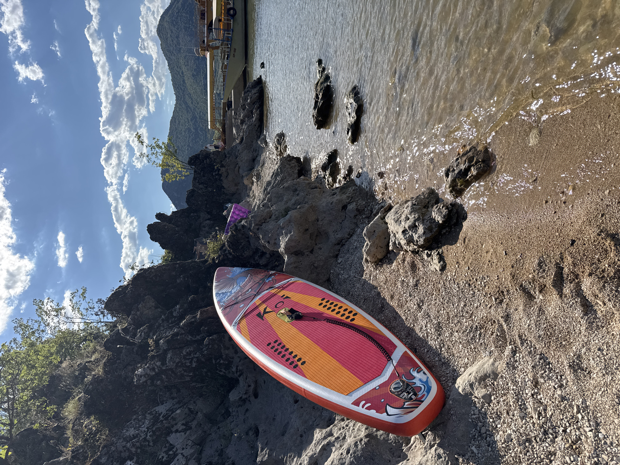 An eye-level, full shot captures a brightly colored stand-up paddleboard leaning against a rocky, pebble-strewn shore on a sunny day at Seoca, Montenegro. The paddleboard is red, pink, and orange with black accents and a graphic design of a bird and waves. A black coiled leash is attached to the board. The water is a murky brown with ripples and reflections of sunlight. Rocks of various sizes are scattered along the shore and in the water. In the background, a light yellow and brown boat with a yellow awning is docked on the right side of the frame. The shore transitions to a rocky cliff face covered in vegetation on the left. Distant green hills and mountains are visible under a bright blue sky with scattered white clouds.