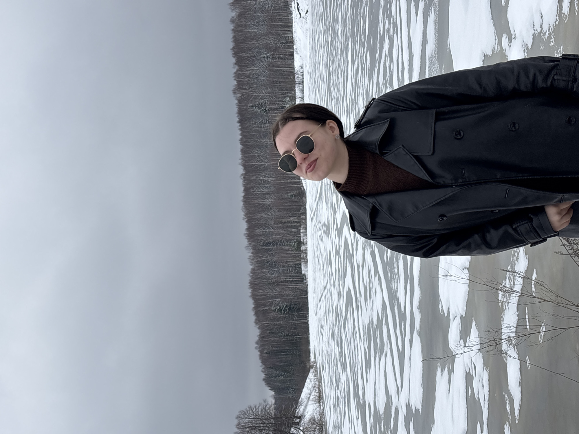 A young woman wearing a brown sweater, black leather trench coat, and round sunglasses stands in front of a partially frozen body of water. Bare trees line the shore. The sky is overcast. The scene appears to be in a natural setting, likely a park or lakeside area in Zasechnyi, Russia.