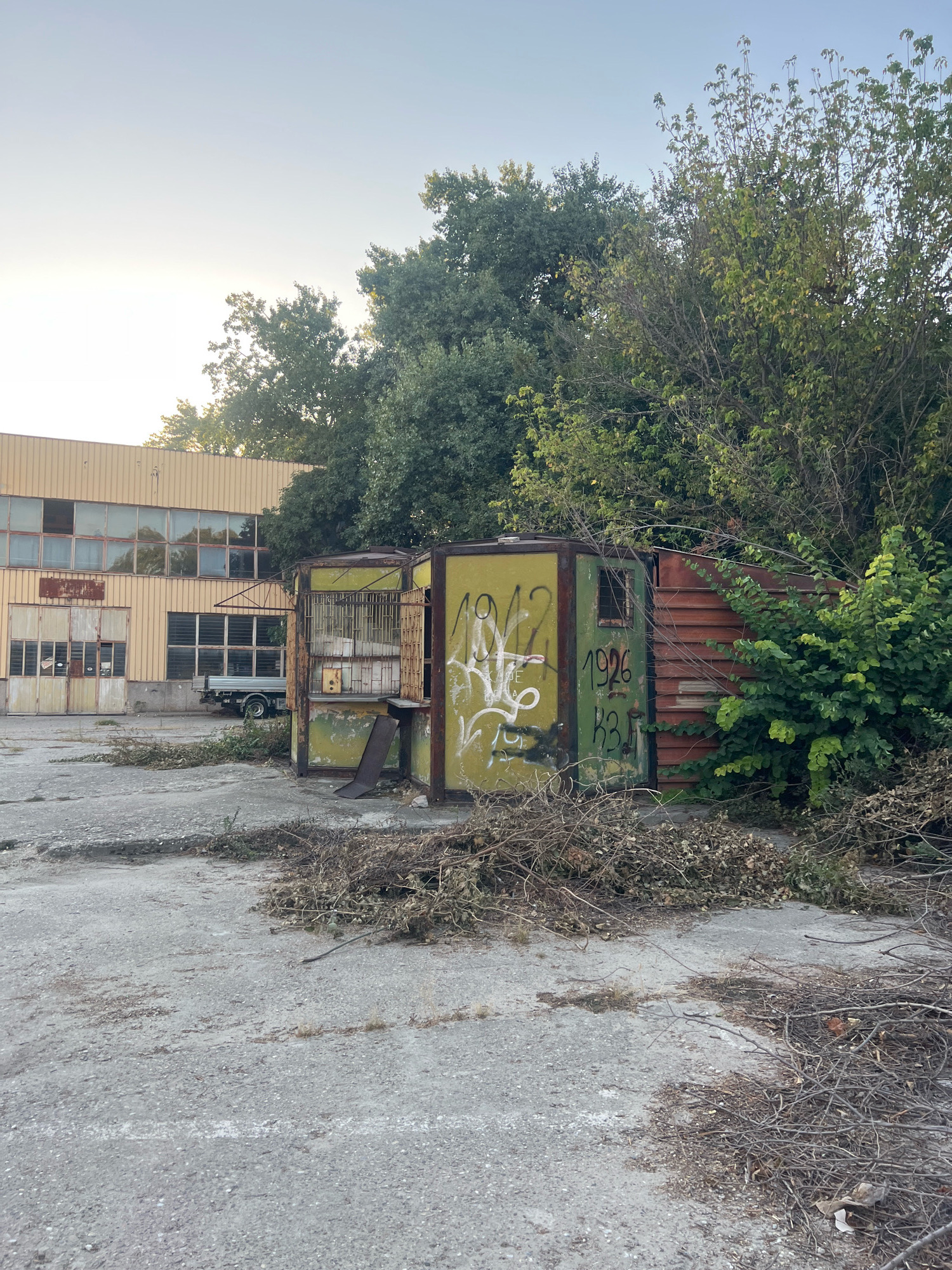 An outdoor industrial lot in Plovdiv, Bulgaria, features a light yellow building with large windows and a flat roof. In front of the building are two small, weathered, yellow and green modular structures, one with a cage-like front. Graffiti, including the numbers "1942" and "1926," is visible on the structures. Piles of dried branches and leaves are scattered on the cracked concrete ground. A small, silver utility truck is parked near the building. Trees and dense foliage surround the area, with a pale, hazy sky overhead.