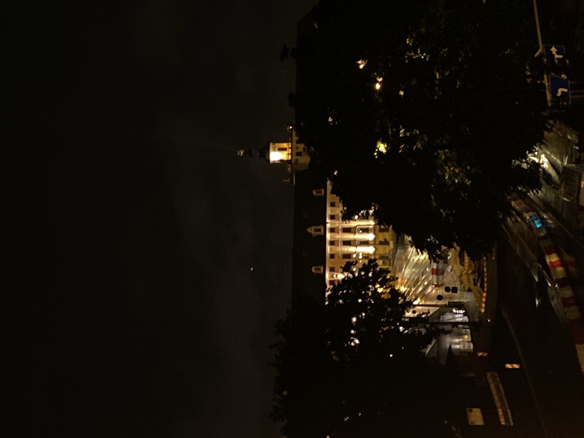 The image depicts a nighttime view of the University of Wrocław (Uniwersytet Wrocławski) in Wrocław, Poland. The university building, illuminated by exterior lights, is visible in the background. A prominent tower with a light at its peak is a focal point of the structure. The foreground shows a dark street with wet pavement, indicating recent rain or damp conditions. There are some trees obscuring parts of the scene. Traffic signs are visible on the right side of the image, and construction barriers with red and white stripes are present on the road. No people are clearly discernible in the image.