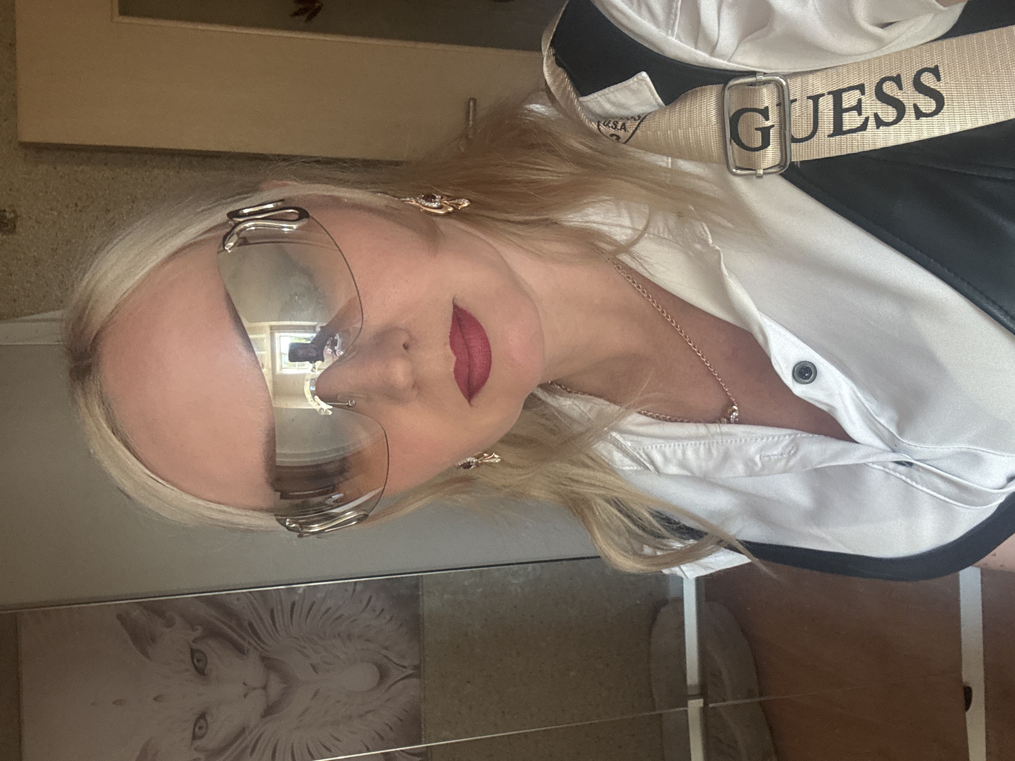 A blonde woman with fair skin and a bright pink lipstick is taking a mirror selfie. She is wearing large, futuristic-style silver sunglasses with reflective lenses that show a reflection of a room with a window and a person holding a phone. She has blonde hair with highlights, and is wearing a white button-down shirt with a black vest over it. A beige strap with the word "GUESS" printed in black letters crosses her chest diagonally. She is also wearing a delicate rose gold necklace with a small pendant and dangling earrings. To her left, a framed artwork of a stylized white cat face is visible on a mirrored surface. The background is a neutral-toned wall with a door handle visible on the right. The lighting is bright, likely from an overhead source, illuminating her face and the surrounding environment. The scene appears to be indoors, in a residential setting. There is no information in the image to indicate the location is Komsomolsk-on-Amur, Russia.