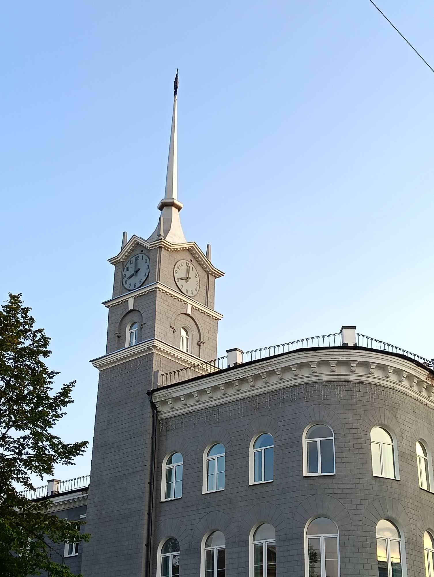 The image shows the Petrozavodsk Railway Station, located in Petrozavodsk, Russia. The building is a grey, multi-story structure with a prominent clock tower topped by a spire. The clock tower has two visible clock faces, each displaying the time. The main body of the building features a curved facade with numerous arched windows, many of which have white frames. A decorative white molding runs along the top of the building, and a black wrought-iron railing encircles a section of the roof. A tree with green leaves is visible on the left side of the image, partially obscuring the building. The sky is clear and blue.
