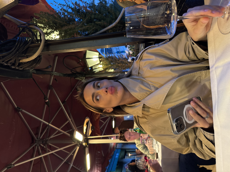 The scene depicts an outdoor dining area in Venice, Italy, observed at dusk or night. A woman with light brown hair, wearing a beige trench coat over a plaid shirt, is seated in the foreground at a table covered with a white tablecloth. She holds a smartphone with a clear case and a circular pop socket in her left hand, on which a silver ring is visible on her ring finger. Her head is turned slightly to the left, and she looks upwards and left with lips intentionally pursed. To her right, a tall-stemmed, empty wine glass is positioned on the table.
Overhead, a red canopy, likely an awning or large umbrella, features multiple attached lights and a visible patio heater. A sturdy black pole with bundled black electrical wires runs vertically on the right side of the frame. In the middle ground, a second woman with dark hair, wearing a light-colored top, is seated at a different table, engaged with a smartphone. A green bottle is on her table. A hand holding a wine glass containing red liquid is partially visible on the far left. Beyond the immediate dining area, a traditional lantern-style street lamp with a white shade illuminates green foliage, likely small trees or bushes. Distant buildings are discernible against a deep blue sky.