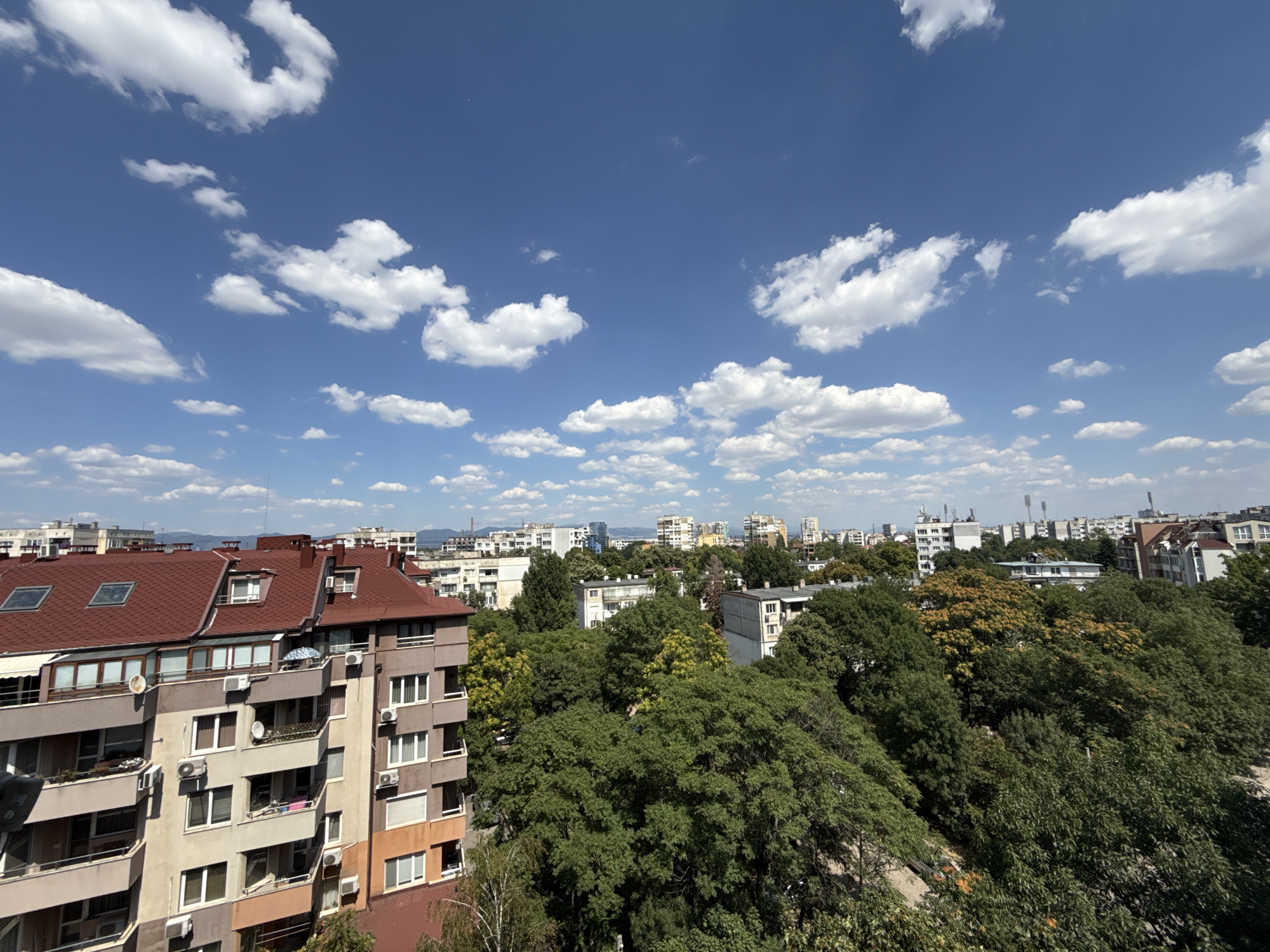 The image shows a view of a city from a high vantage point, likely an apartment building, with a clear blue sky dotted with fluffy white clouds. In the foreground, a multi-story residential building with a reddish-brown tiled roof is visible. The building has numerous windows and balconies, some with air conditioning units attached. Adjacent to this building, to the right, is a dense cluster of green trees, some of which show hints of yellowing foliage, suggesting a late summer or early autumn season. Beyond the trees, the cityscape unfolds with more residential buildings of varying heights and architectural styles, predominantly in white and light colors, with some appearing to be older Soviet-era constructions. In the distance, the outlines of more buildings and possibly hills or mountains can be seen against the horizon. The scene is indicative of a residential area within a city, likely Sofia, Bulgaria, given the architectural style of the buildings and the climate suggested by the greenery and sky. There are no people or specific actions depicted in the image, making it a static environmental portrait of an urban landscape.