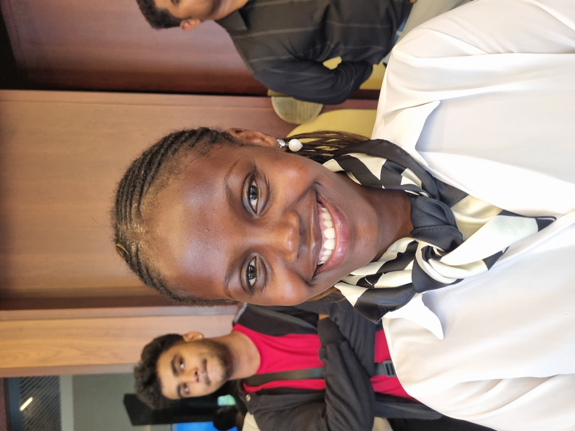A medium-shot, rotated image shows a woman smiling broadly at the camera. She has dark skin, dark eyes, and braided hair with a light shimmer. She is wearing a white blouse and a black-and-white scarf. She is wearing a pearl earring.
In the background are two men with darker skin and dark hair. The first is wearing a red shirt and black jacket, and the second is wearing a black striped shirt. The background shows elements of an interior space.
There is no evidence in the image that allows for the determination of a specific location, city, or country. Thus, it is impossible to determine if this image was taken in Kottukkara, India.
