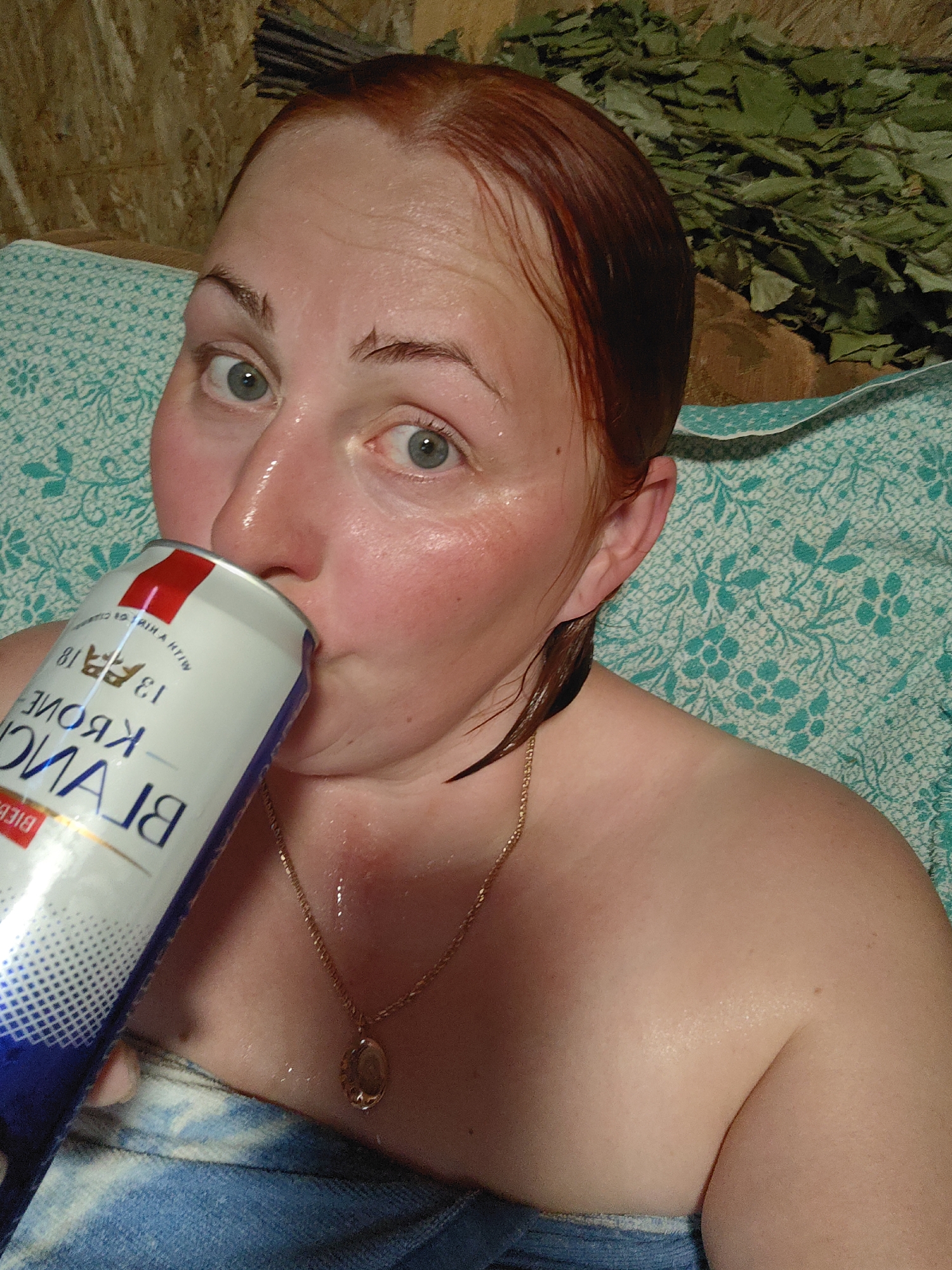 A red-haired woman with damp skin is drinking from a can labeled "KRONE BLANC" in what appears to be a sauna. She has a gold necklace with a pendant. A green patterned cloth is draped around her. Behind her is a bundle of birch leaves. The location is Фоки, Russia.