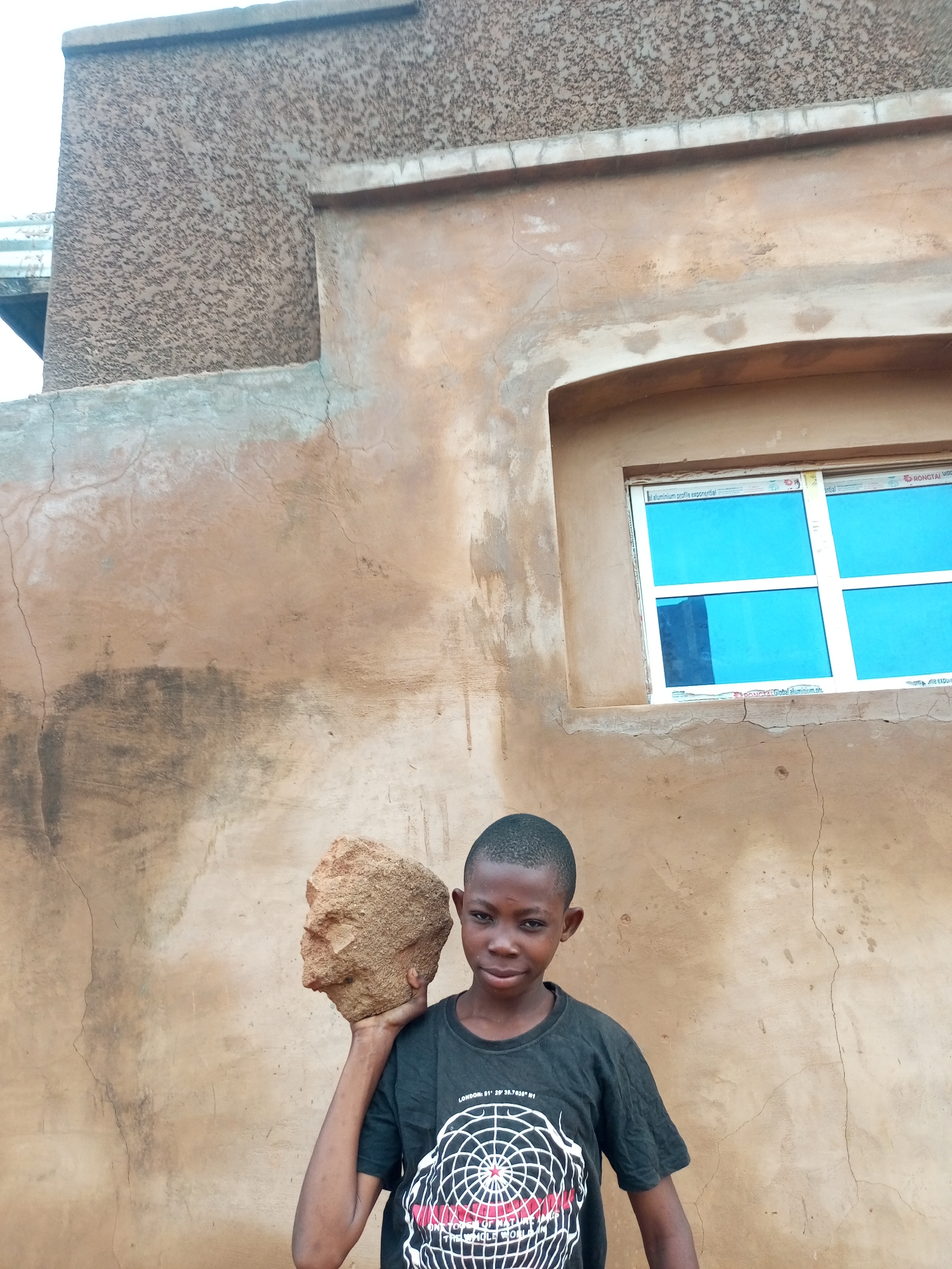 A young boy with short dark hair and brown skin stands in front of a textured, tan-colored wall. He is wearing a black t-shirt with a white circular graphic. The boy is holding a large, rough, reddish-brown rock on his right shoulder with his right hand. To the boy's right, there is a rectangular window with blue panes framed in white. The wall has areas of cracking and discoloration. The scene appears to be outdoors, in front of a building.