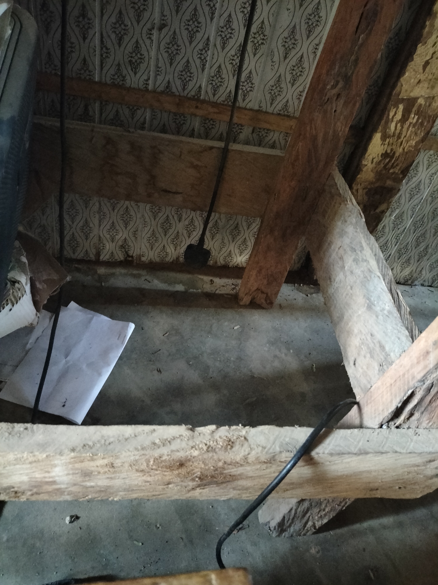 The image depicts the interior of what appears to be an attic or storage space in Jalingo, Nigeria. The scene is characterized by exposed wooden beams forming the roof structure, with some of the wood showing signs of age and wear. The ceiling is covered with patterned wallpaper, some sections of which appear to be peeling. The floor is concrete and dusty, with scattered debris.
In the foreground, a thick wooden beam runs horizontally, with a black cable resting on it. Another black cable snakes across the scene, disappearing behind a piece of wood. To the left, a piece of white paper is crumpled on the floor. In the background, a black shovel is propped against the wallpapered wall. The lighting is dim, suggesting natural light entering from an unseen source or artificial lighting that is not prominently visible. The overall impression is one of a neglected or underutilized space.