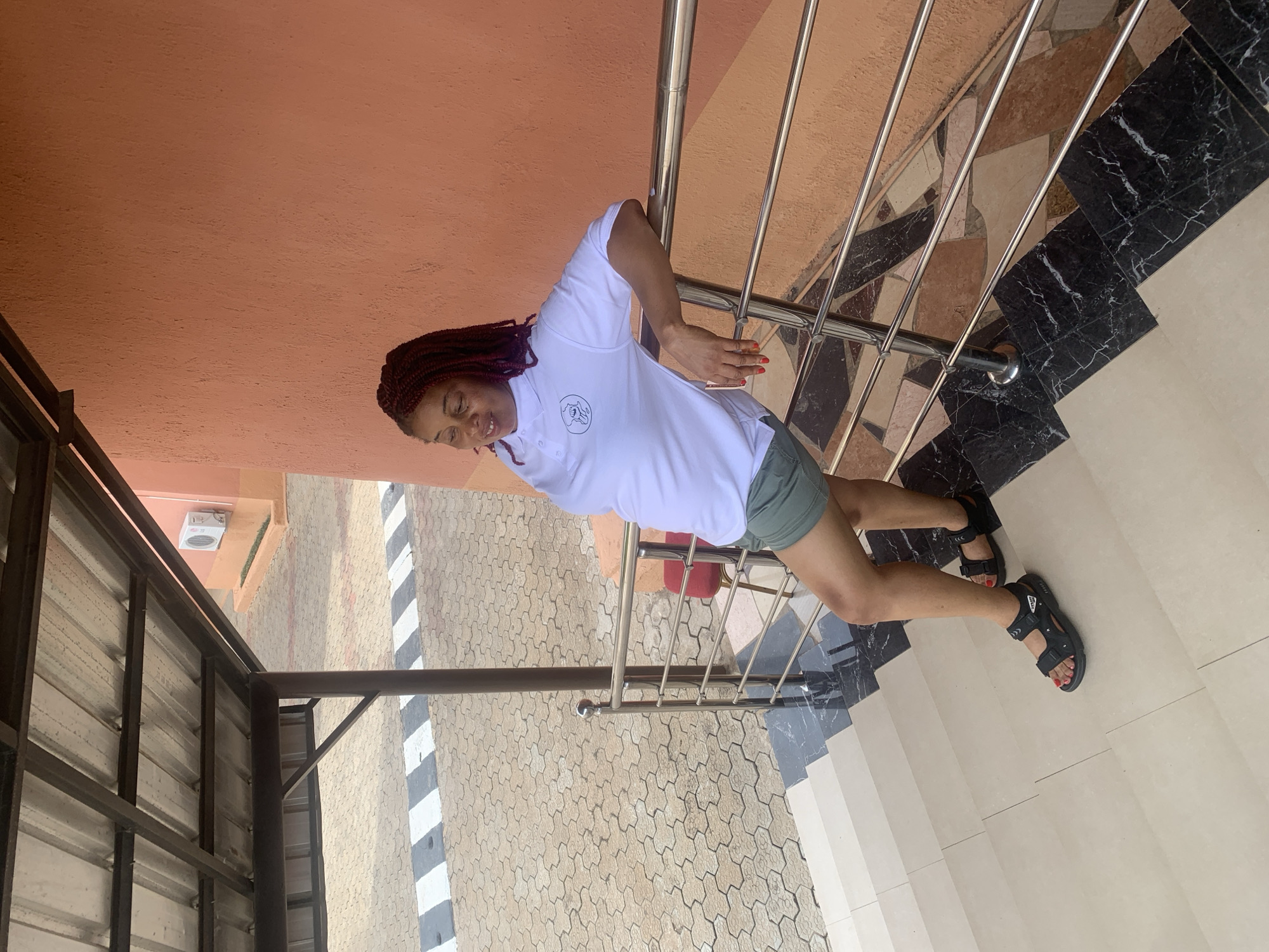 A woman stands on the steps of a building in Gura Top, Nigeria. She holds onto a metal railing. She wears a white, short-sleeved collared shirt, and green shorts. She has braided red hair. She wears black sandals. The building features orange walls, and the steps are a mix of black marble and white tile. A paved area with black and white markings is visible outside.