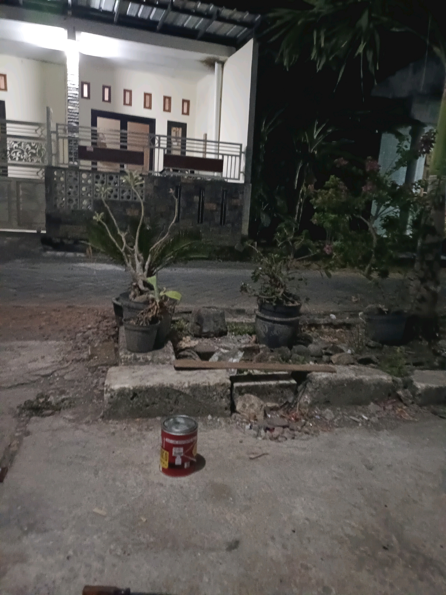 This media file captures a nighttime scene in Rancang Kencono, Indonesia. The primary focus is a residential area with a house partially visible in the background. The house features a white facade, multiple rectangular windows, and a metallic roof. A fence with decorative elements separates the property from the street.
In the foreground, the scene is dominated by the street and a sidewalk area. Several potted plants are placed along the roadside, adding a touch of greenery to the otherwise concrete and asphalt environment. One plant in a dark pot on a concrete ledge is particularly prominent, with thin branches and some sparse leaves. Another larger pot is visible further back, also containing a plant.
A bright red cylindrical object, likely a can, rests on the concrete ground in the lower right quadrant of the image. The can has some text and graphics on its label, but it's difficult to decipher clearly due to the image quality and lighting. The overall impression is one of a quiet, everyday scene in a residential neighborhood at night. The lighting suggests artificial light sources, possibly from the house or streetlights, casting shadows and creating a somewhat muted ambiance. The presence of potted plants and the architectural style of the house provide subtle clues about the location.
