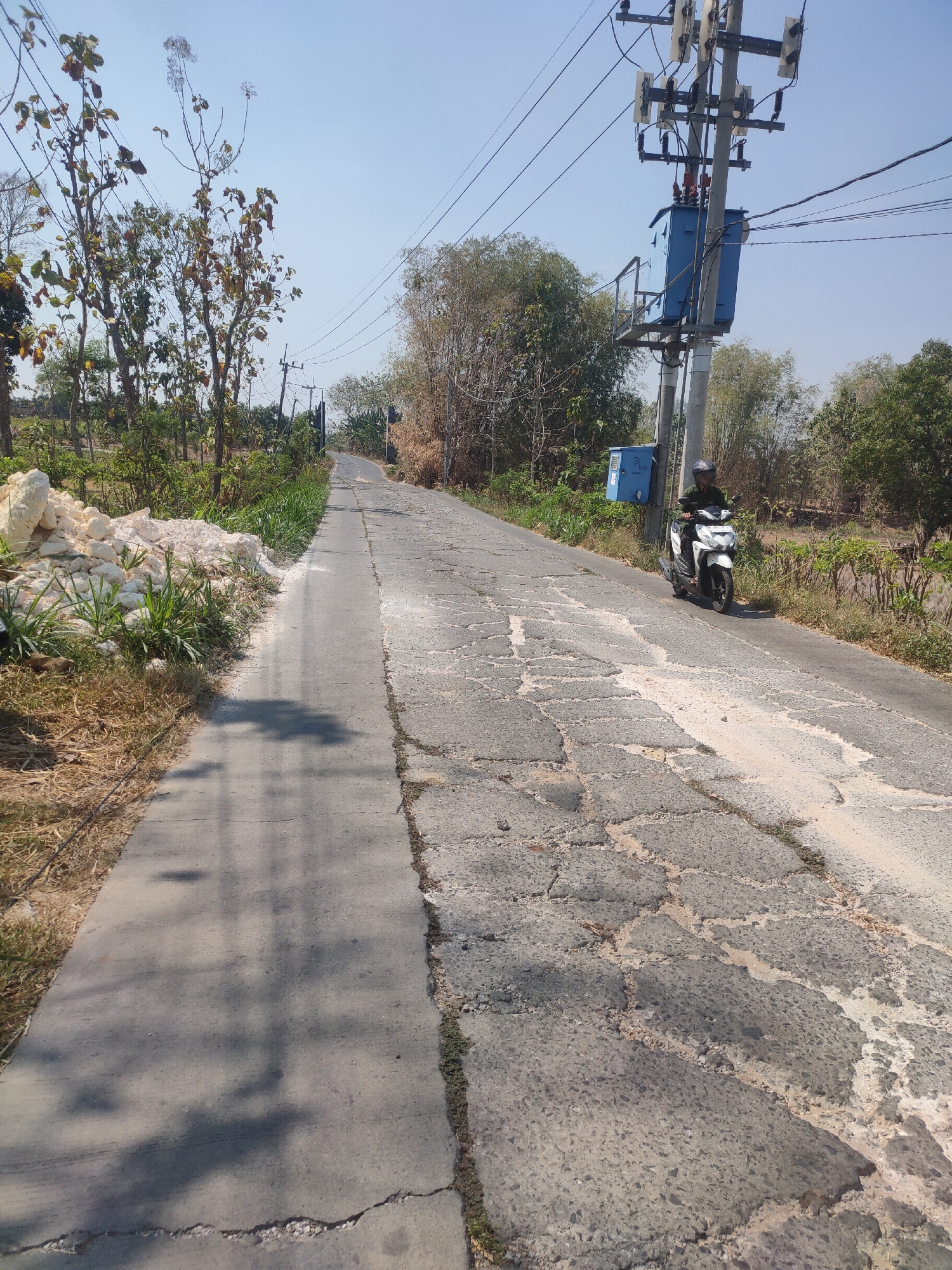 In Bluri, Indonesia, a man rides a white scooter on a cracked asphalt road. The road has a separate, smoother concrete path on the left side. Power lines and a utility pole with a blue transformer are visible on the right side of the road. The surrounding environment is rural, with trees, dry grass, and a pile of white material, possibly limestone, on the left side of the road. The sky is clear and blue.