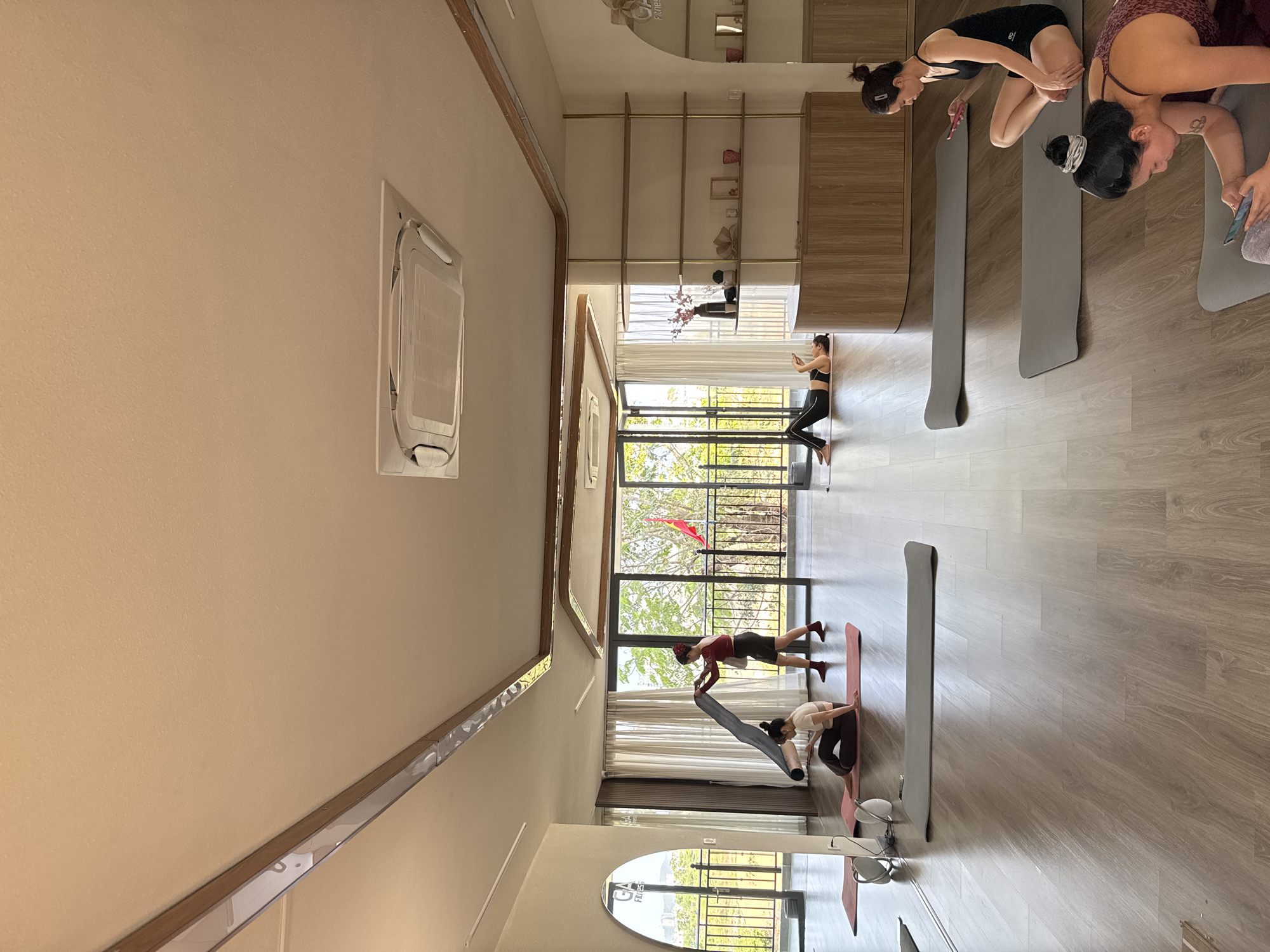 A group of people are engaged in yoga within a studio setting, the city being Unknown, Unknown. The studio has wooden flooring, large windows allowing natural light, and built-in shelving along one wall. Several individuals are on yoga mats, performing different poses, while some are standing and stretching. Some people are looking at their phones. The environment is well-lit and seems to be designed for fitness activities.