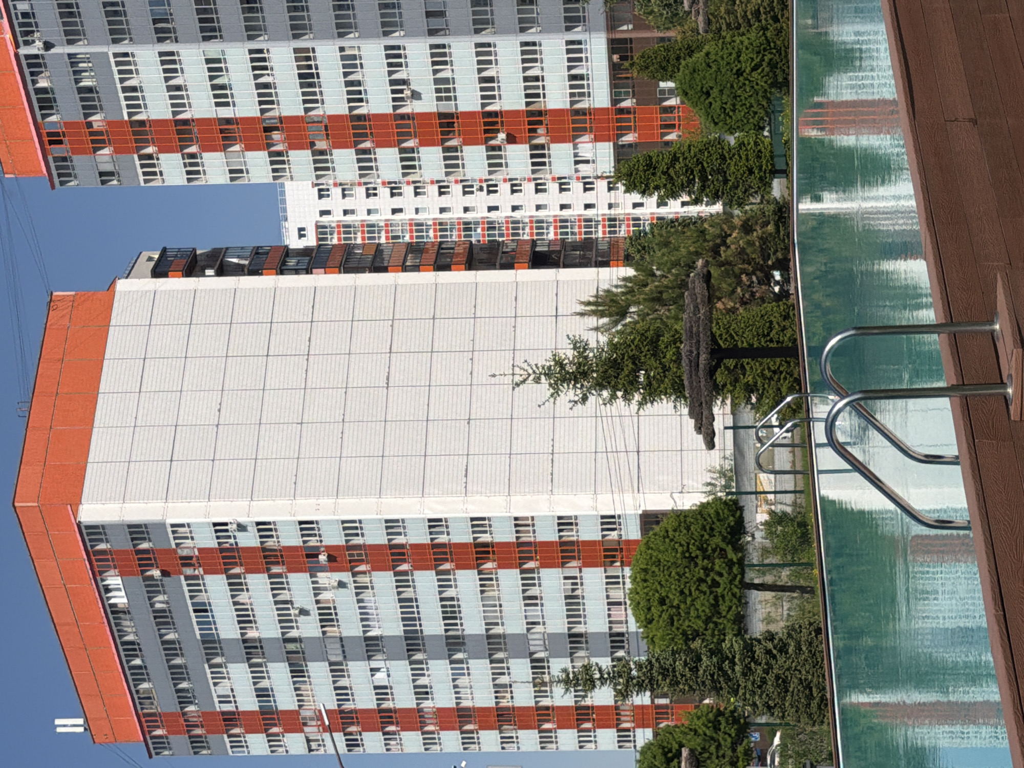 The image displays an urban scene in Krasnoyarsk, Russia, featuring tall residential buildings alongside a recreational swimming pool. Two prominent, multi-story apartment blocks dominate the left side of the frame, characterized by repeating patterns of light-colored panels and numerous rectangular windows, with distinctive reddish-orange horizontal stripes across their facades. A third, smaller building is visible in the background between these two structures.
To the right, a rectangular swimming pool with clear, blue-green water is bordered by a dark brown wooden deck. Stainless steel handrails are visible, providing access to the water. The pool's surface reflects the surrounding buildings and the bright, clear blue sky.
Various green trees and bushes are scattered throughout the scene, particularly around the base of the buildings and along the pool area. The bright, clear sky suggests sunny weather, likely during midday or early afternoon. No people are visible, and no specific activity is taking place, creating a calm urban landscape. No visible text is present in the image.