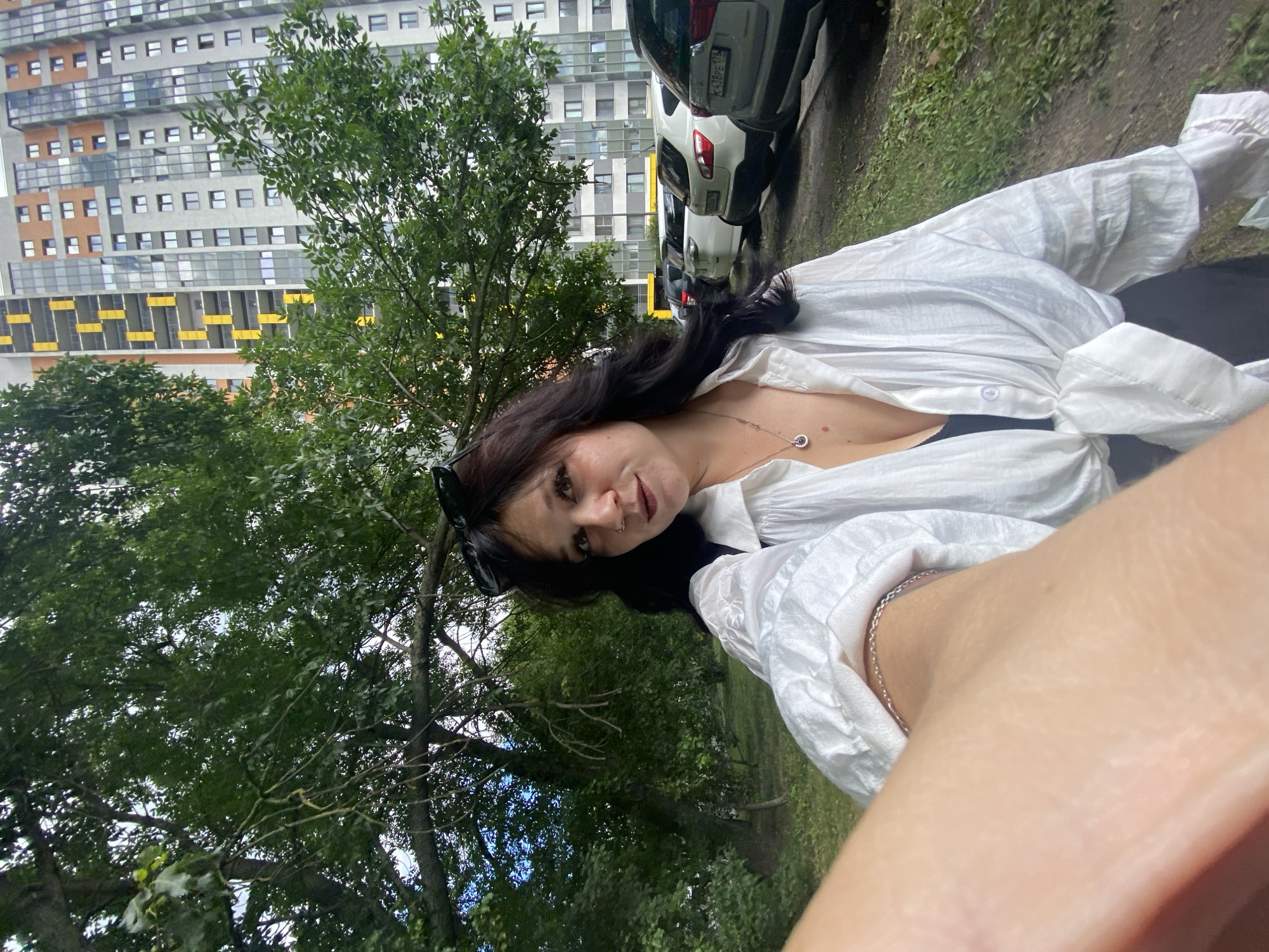 An outdoor, medium-shot, selfie-style image shows a young adult, appearing to be female, with long, dark, wavy hair and wearing a white oversized button-up shirt tied at the waist over a dark top, and black pants. She has sunglasses perched on her head and is wearing a pendant necklace and a nose ring. Her left arm and hand are in the foreground, partially obscuring the view. The background includes a large tree with green foliage and a modern, high-rise apartment building with balconies and yellow accent panels. Several cars are parked in a lot behind the tree. The scene appears to be a residential area in Saint Petersburg, Russia. The lighting suggests an overcast day.