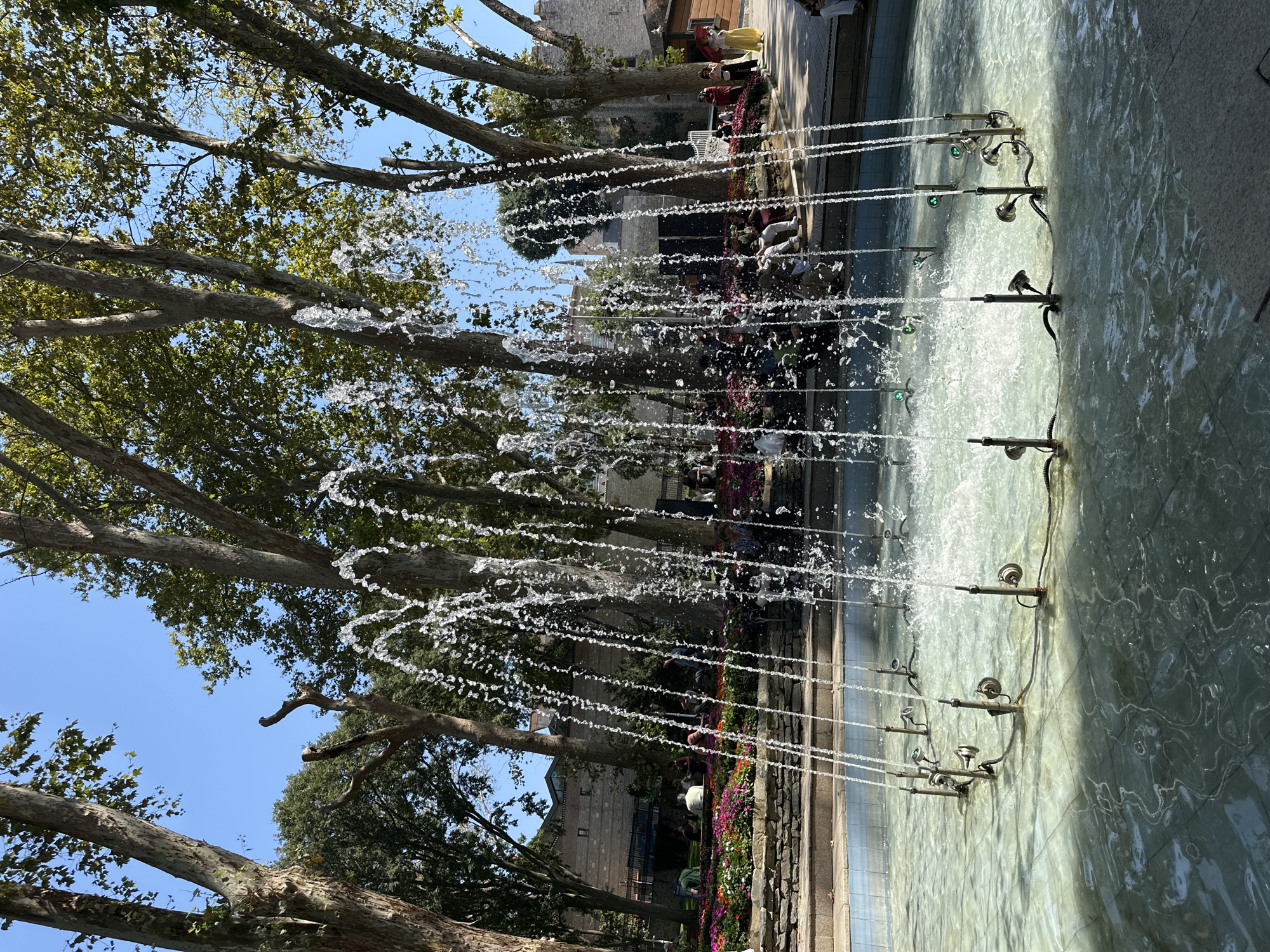 This image captures a water fountain in a public park, likely in Istanbul, Turkey. The fountain is circular, with multiple jets of water spraying upwards and outwards, creating a shimmering effect. The water is clear and shallow, revealing the tiled bottom of the basin. Surrounding the fountain, there are lush flower beds in vibrant shades of pink, red, and white, bordered by stone walls. Tall, mature trees with broad leaves provide shade, their branches reaching towards the clear blue sky. In the background, several people are seated and standing, some observing the fountain and the colorful flowers. The architecture in the far background, with its stone walls and a distant building, suggests a historical or cultural context, consistent with many public spaces in Istanbul. The scene is bathed in bright sunlight, indicating a warm, pleasant day.