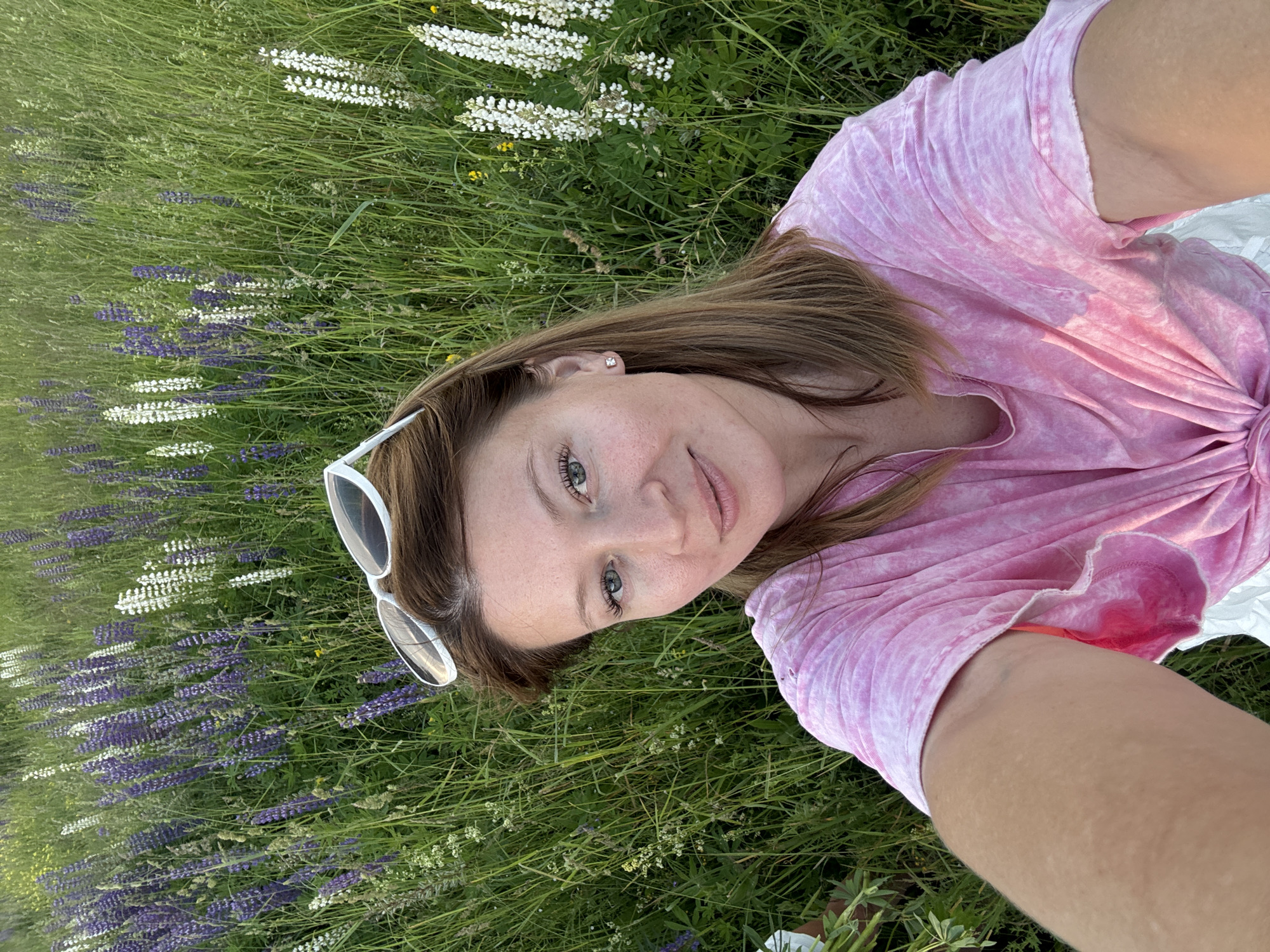 A selfie is taken by a fair-skinned woman with medium-length brown hair and blue eyes, wearing a pink tie-dye t-shirt and white sunglasses perched on her head. She is outdoors, in a field of tall green grass dotted with purple and white lupine flowers, suggestive of a rural or parkland setting in Vassyata, Russia. The lighting indicates it is daytime, possibly late afternoon or early evening, given the warm glow. The woman is smiling and looking directly at the camera. The background consists of a dense field of flowers and vegetation, with no urban structures visible.