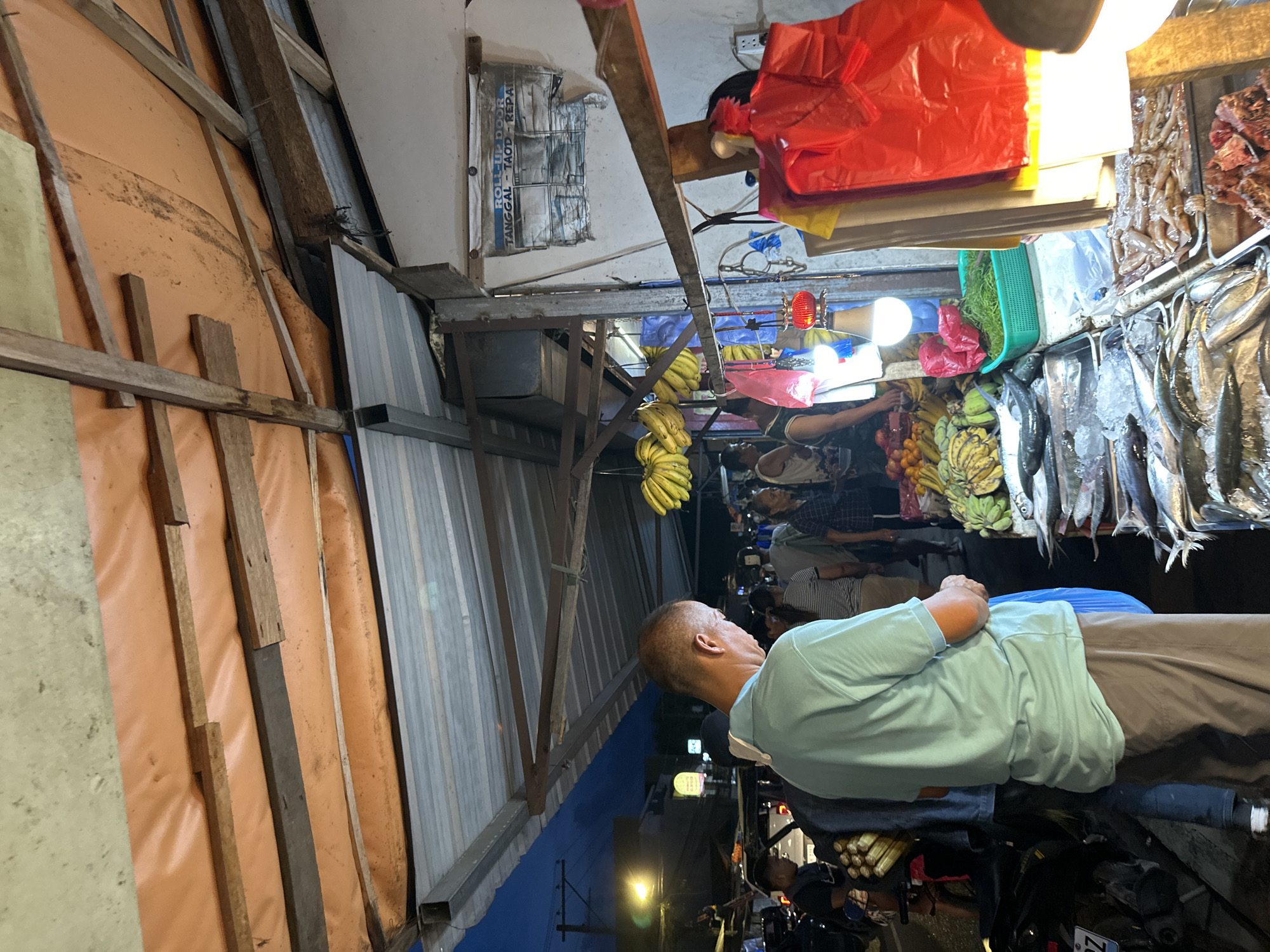 The image depicts a bustling outdoor market scene at dusk, possibly in Davao City, Philippines. A man in a light blue polo shirt and brown pants is the primary subject in the foreground, facing away from the camera and looking towards a stall. He is holding a blue plastic bag. The market stall is laden with fresh produce, including bananas, and an array of seafood displayed on ice, such as fish and what appears to be shrimp. Several other individuals are visible in the background, navigating through the market. The stall is covered by a corrugated metal roof supported by wooden beams, with an orange tarp overhead. A sign reading "ROLL-UP DOOR TANGGAL - TAOD - REPAIR" is affixed to a white wall behind the stall, suggesting services offered in the vicinity. Lighting appears to be a mix of overhead bulbs and ambient twilight, contributing to the overall atmosphere of a lively marketplace.