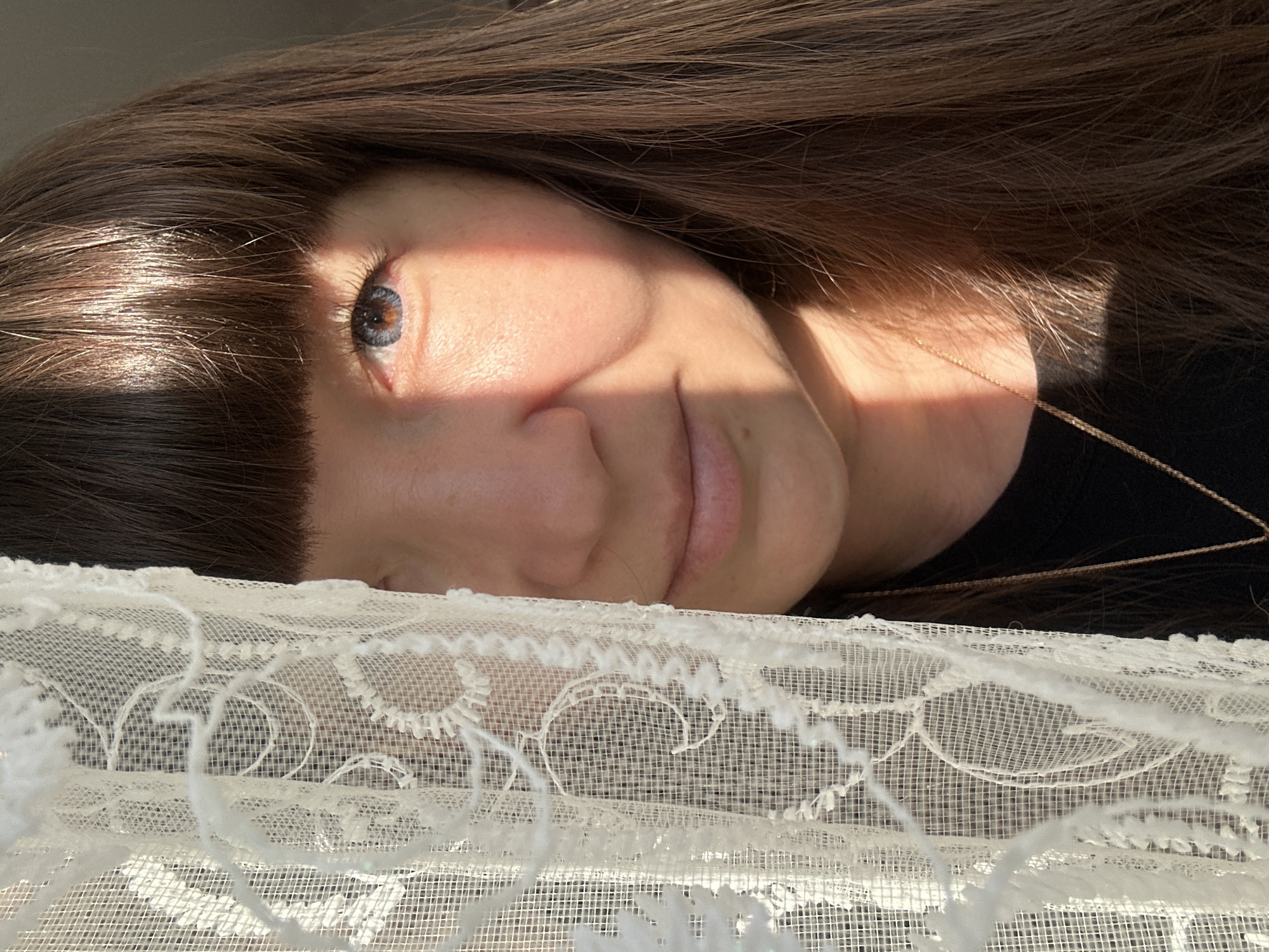 The image is a close-up portrait of a woman partially obscured by white lace. She has long, dark brown hair with bangs and a single visible blue eye. The sunlight casts a shadow across her face, highlighting the texture of her skin. She is wearing a black shirt and a delicate gold chain. The background is out of focus, but suggests an indoor setting. The photographer is likely in Chaikovsky, Russia, as indicated by the context provided.