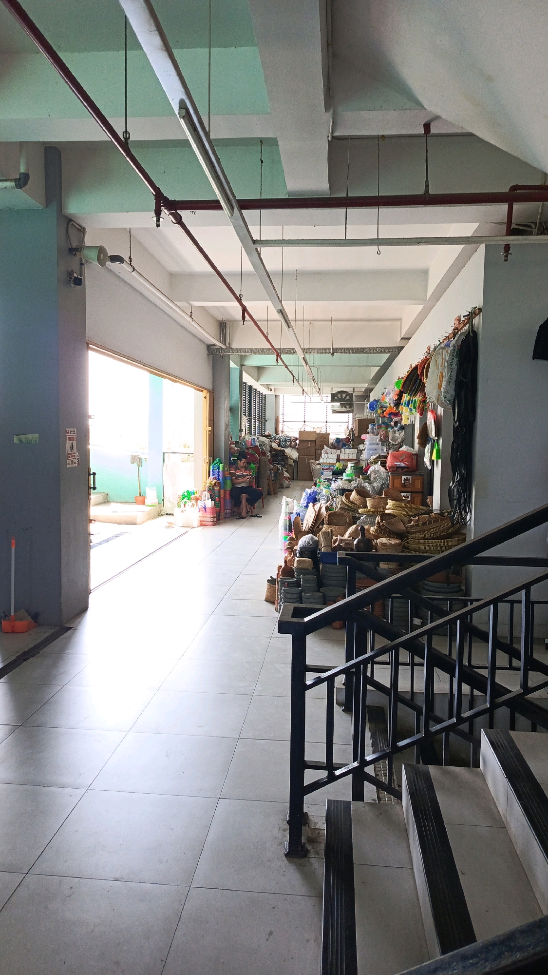 The image depicts the interior of a store or warehouse in Ponorogo, Indonesia, likely a retail space selling various household goods and crafts. The scene is viewed from a slightly elevated perspective, looking down a long corridor with an open doorway on the left and a staircase on the right.
The environment appears to be an indoor, covered walkway or hallway with a tiled floor and a high ceiling. Exposed pipes and fluorescent lighting fixtures are visible along the ceiling. The walls are painted in neutral tones.
On the left side, the open doorway reveals a brightly lit exterior area, possibly a loading dock or an open-air market stall. Inside the store area, a variety of items are neatly arranged and stacked along the right wall and down the corridor. These include woven baskets, bags, colorful plastic containers, and what appear to be other craft or household items. Some boxes are stacked in the background. A man is seated on a chair in the middle ground on the left, appearing to be a shopkeeper or employee.
The foreground features a black metal staircase with wooden steps, indicating a possible mezzanine or upper level of the store. The overall impression is one of a busy, functional retail or storage space, characteristic of market areas in Indonesian cities.