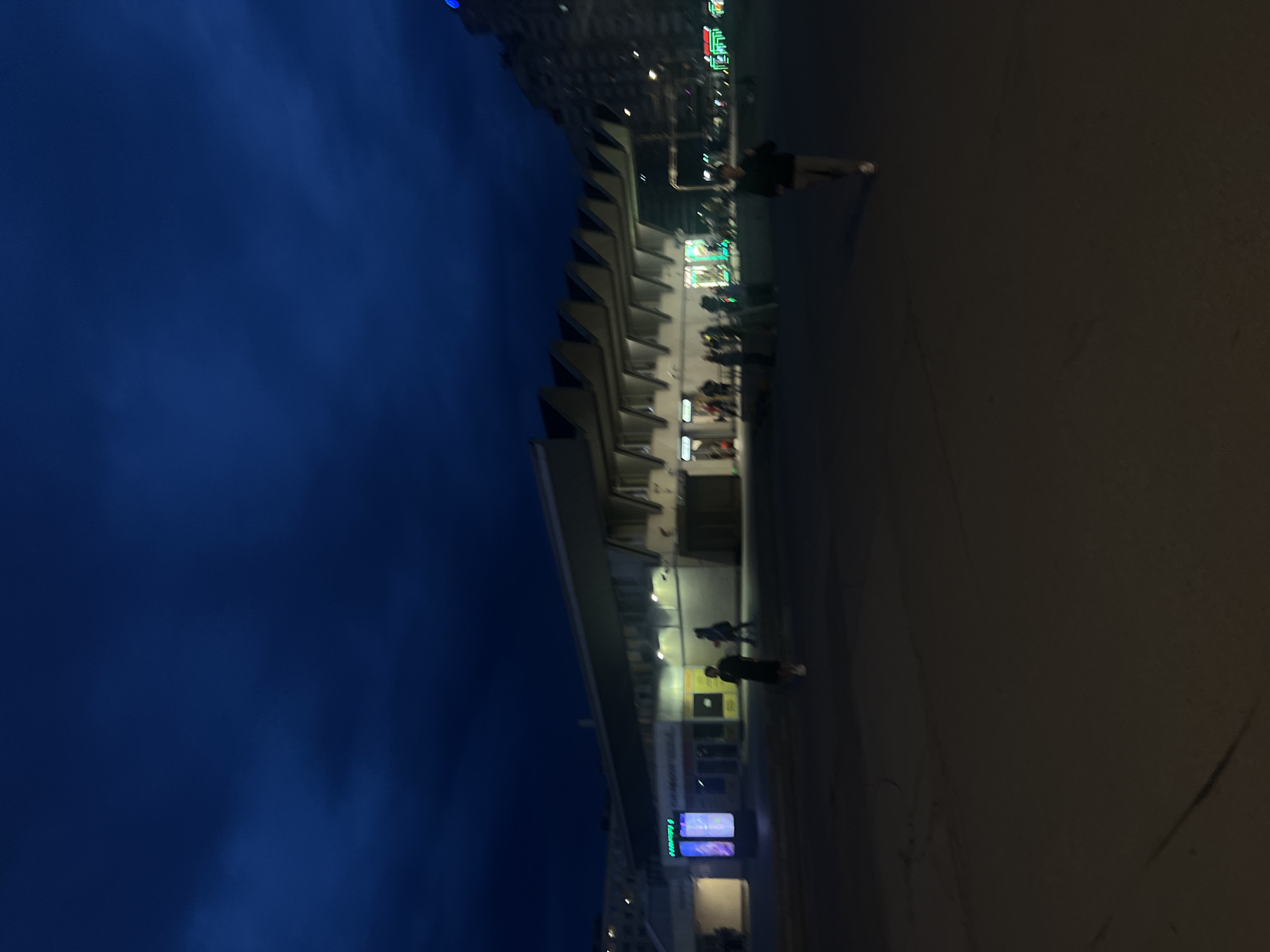 The image depicts a nighttime urban scene in Saint Petersburg, Russia, featuring the Oktyabrsky Grand Concert Hall. The deep blue sky is overcast with dark clouds. In the foreground, a large, dark paved area stretches across the lower portion of the frame.
To the left, a multi-story building with illuminated windows is partially visible. Adjacent to it, a tall, vertical digital display or advertisement board, illuminated with blue and purple tones, stands upright. A lone individual in dark clothing and light shoes walks from left to right past this display. Further left and slightly behind, another person in dark attire walks away from the viewer.
The central and most prominent structure is the Oktyabrsky Grand Concert Hall (BKZ Oktyabrsky), identifiable by its distinctive angular, zig-zag roofline and broad facade. The building's lower levels feature multiple large windows, some emanating warm yellow and green light, suggesting interior illumination. Several indistinct figures are visible near the building's entrances and along its illuminated sections.
To the right of the concert hall, a tall, multi-story building with a repetitive facade rises into the dark background. A single bright blue light, likely an illuminated sign or architectural detail, is visible near its peak. In the mid-ground, walking towards the right foreground, is an individual in a dark top and light-colored pants. The overall scene is lit primarily by artificial sources from the buildings and streetlights, with the entire image appearing slightly out of focus.