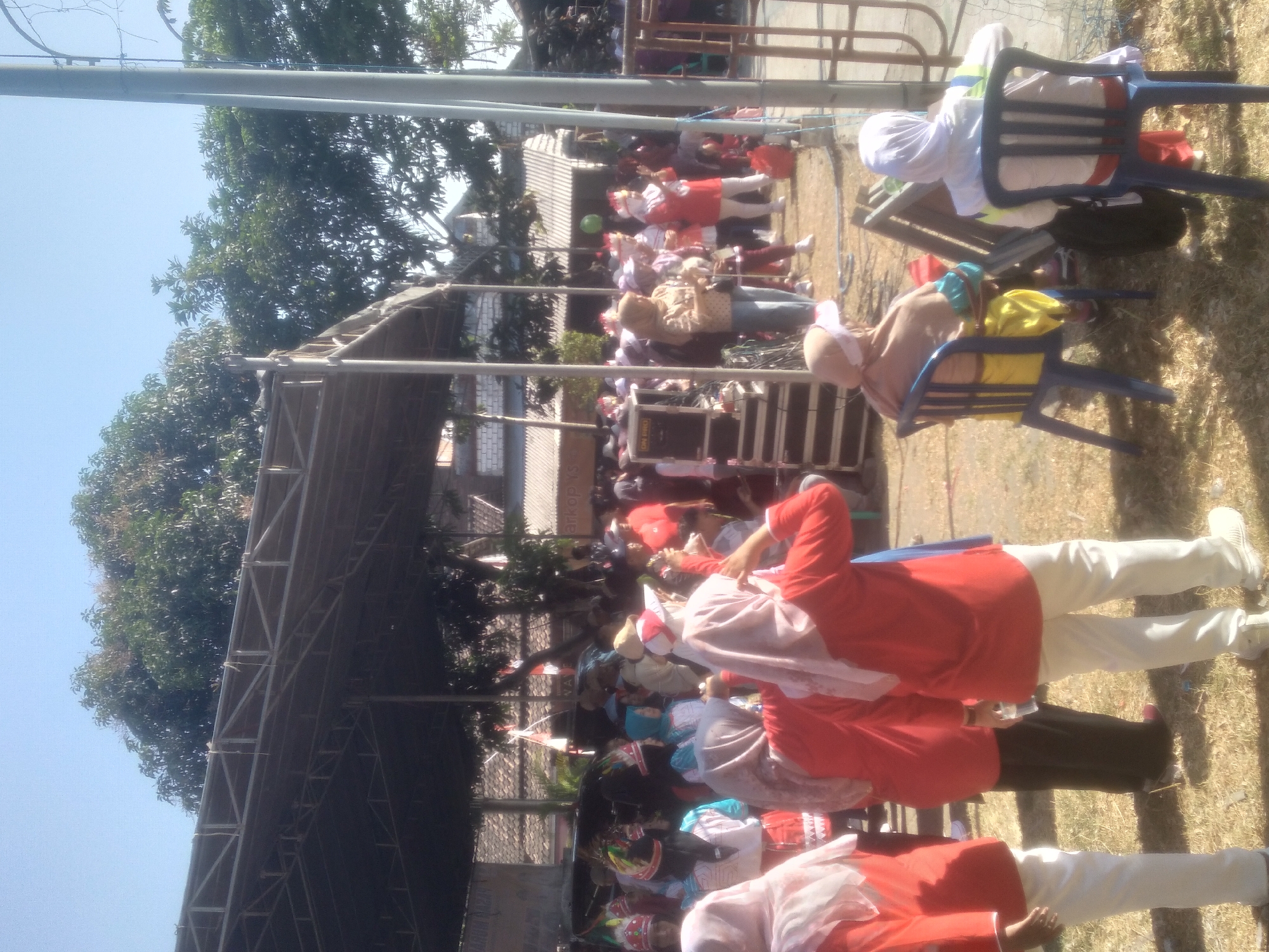 In the city of Lamongan, Indonesia, a group of people, primarily women and children, are gathered outdoors under a shaded structure, possibly for an event or festival. The weather appears to be sunny and clear.
In the foreground, several individuals are visible from behind. One woman in a red top and white pants is standing, facing away from the camera, with her right hand raised to her head, possibly adjusting her headscarf. To her left, another person in a red top and black pants is also standing. Further to the left, a person wearing a white headscarf and a red top is seen from the side.
In the mid-ground, a crowd of people are milling around. Some are wearing colorful headwear, suggesting participation in a parade or performance. Others are observing the scene. There is a stage or platform area with sound equipment visible on the left side of the image. A blue plastic chair is occupied by a person wearing a light brown headscarf and a yellow garment. Another blue plastic chair is occupied by a person in a white headscarf and a red and white top.
The background shows trees and buildings with tiled roofs, typical of Indonesian architecture. A tall, slender pole, possibly a light post or flagpole, stands prominently on the right side of the frame. The ground is covered with dry grass and scattered debris. The overall scene suggests a public gathering in a semi-rural or suburban setting.