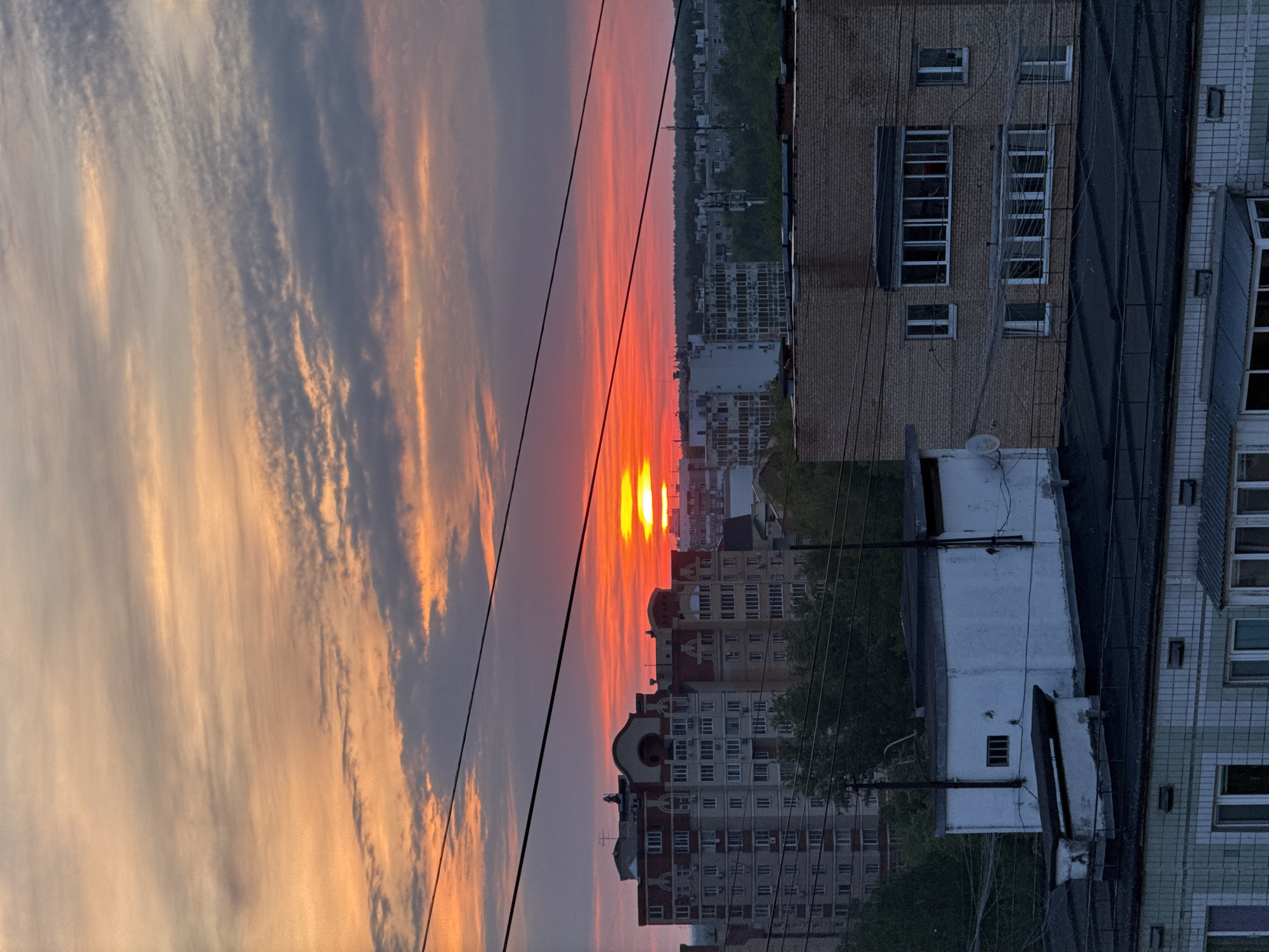 The image shows a sunset over the city of Dolgoprudny, Russia. The sky is filled with clouds, colored with orange and red hues from the setting sun. The sun is positioned low in the horizon, with light reflecting off of the city's buildings. Various buildings of different heights and architectural styles are scattered across the cityscape. Power lines stretch across the foreground. A hill with trees is on the horizon.