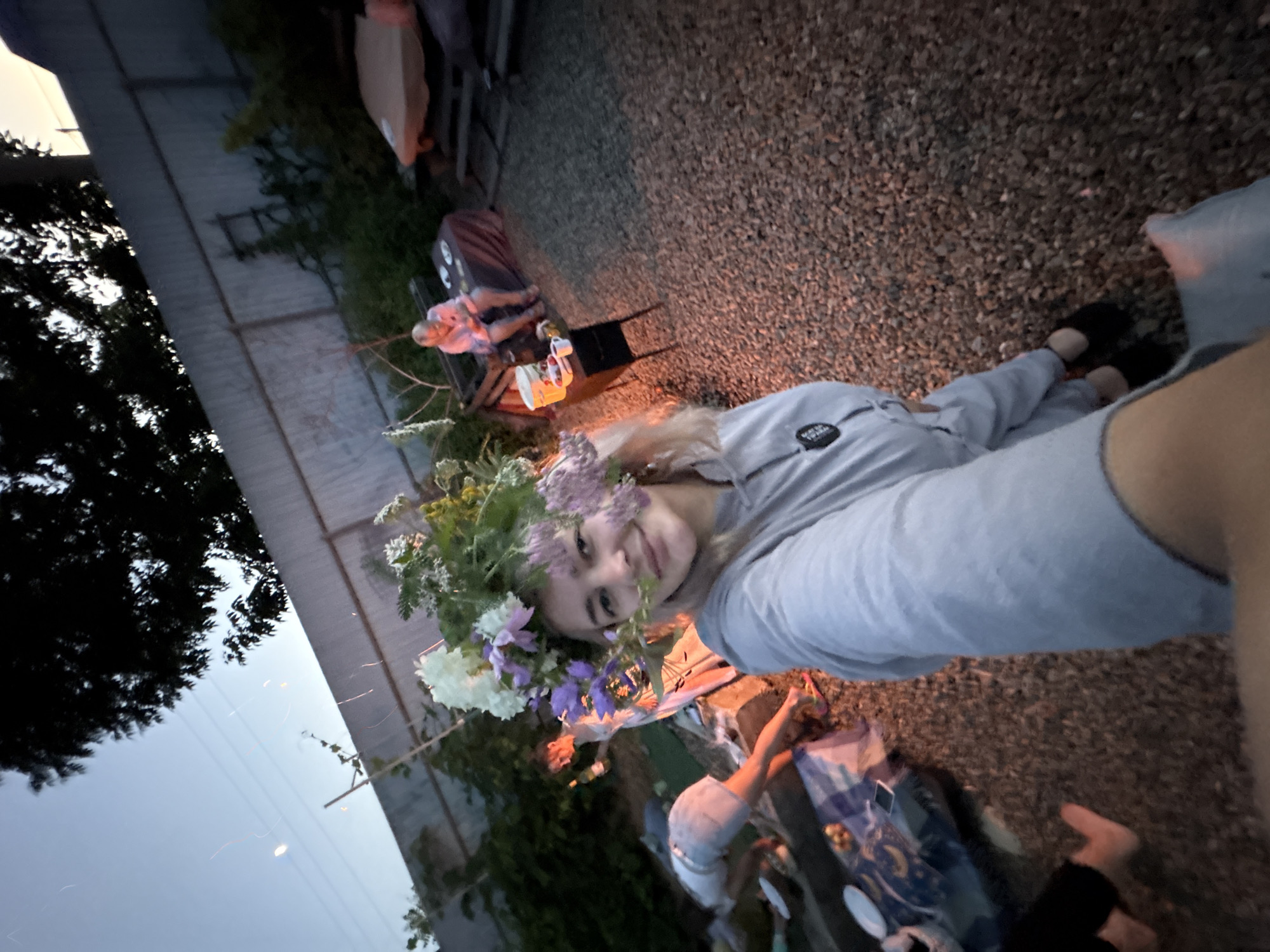 In Birobidzhan, Russia, a young woman with a floral wreath on her head is taking a selfie outdoors at dusk. She is wearing a grey hoodie. In the background, several people are gathered on the ground. One person is sitting at a table with food, another is leaning over a picnic blanket, and others are partially visible. The area appears to be a backyard or outdoor gathering space with gravel on the ground and a wooden fence in the background. The lighting suggests it is evening, with some ambient light from a nearby lamp or fire.